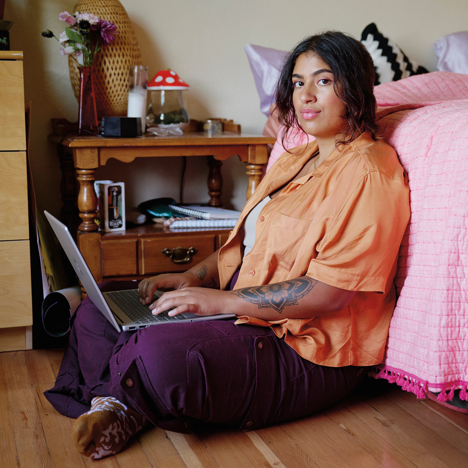 Een persoon zit op de grond met een laptop. Ze draagt een oranje shirt en een paarse broek. Een bed en meubels op de achtergrond.