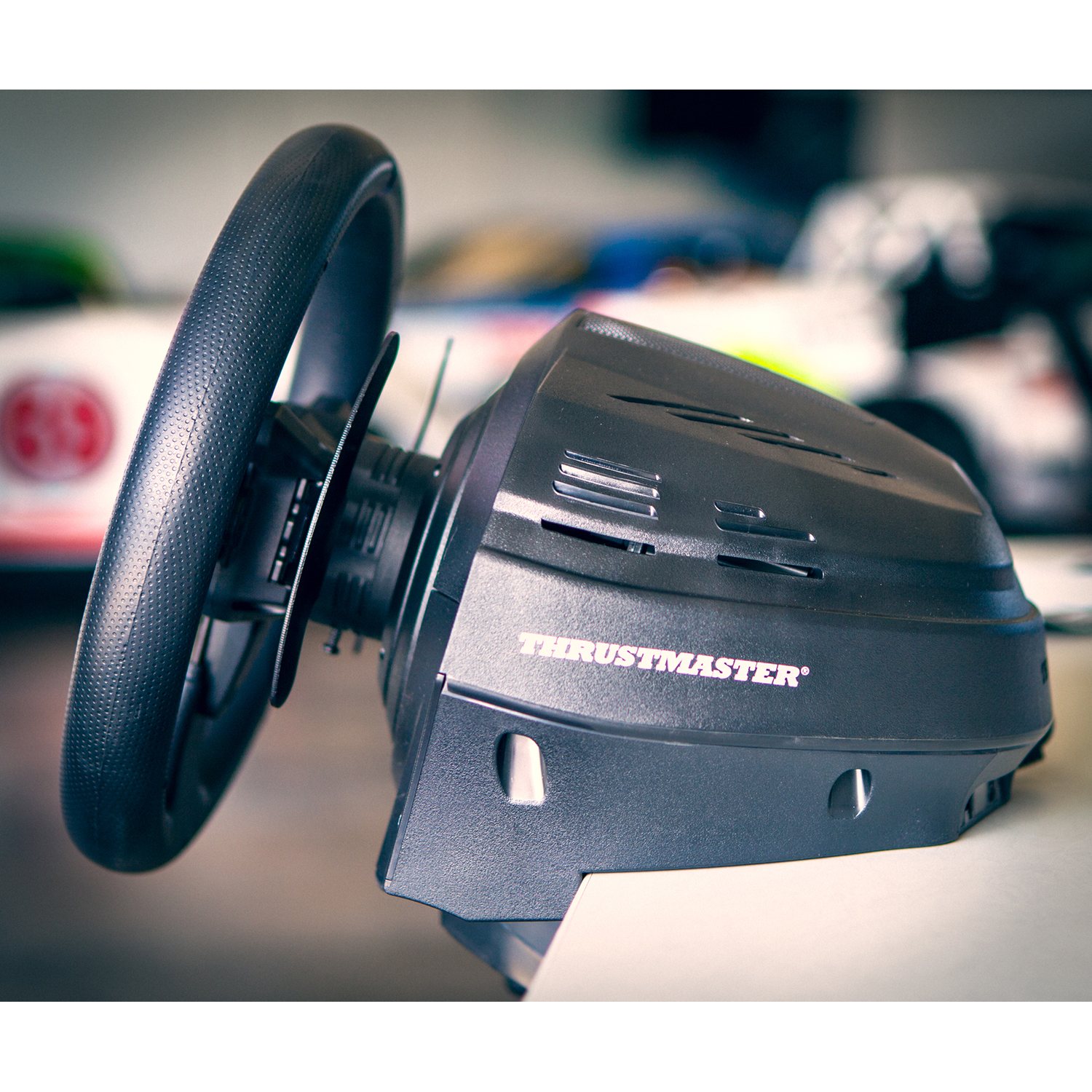 Een zwart Thrustmaster racestuur, met de merknaam zichtbaar, op een licht oppervlak. De achtergrond is wazig.
