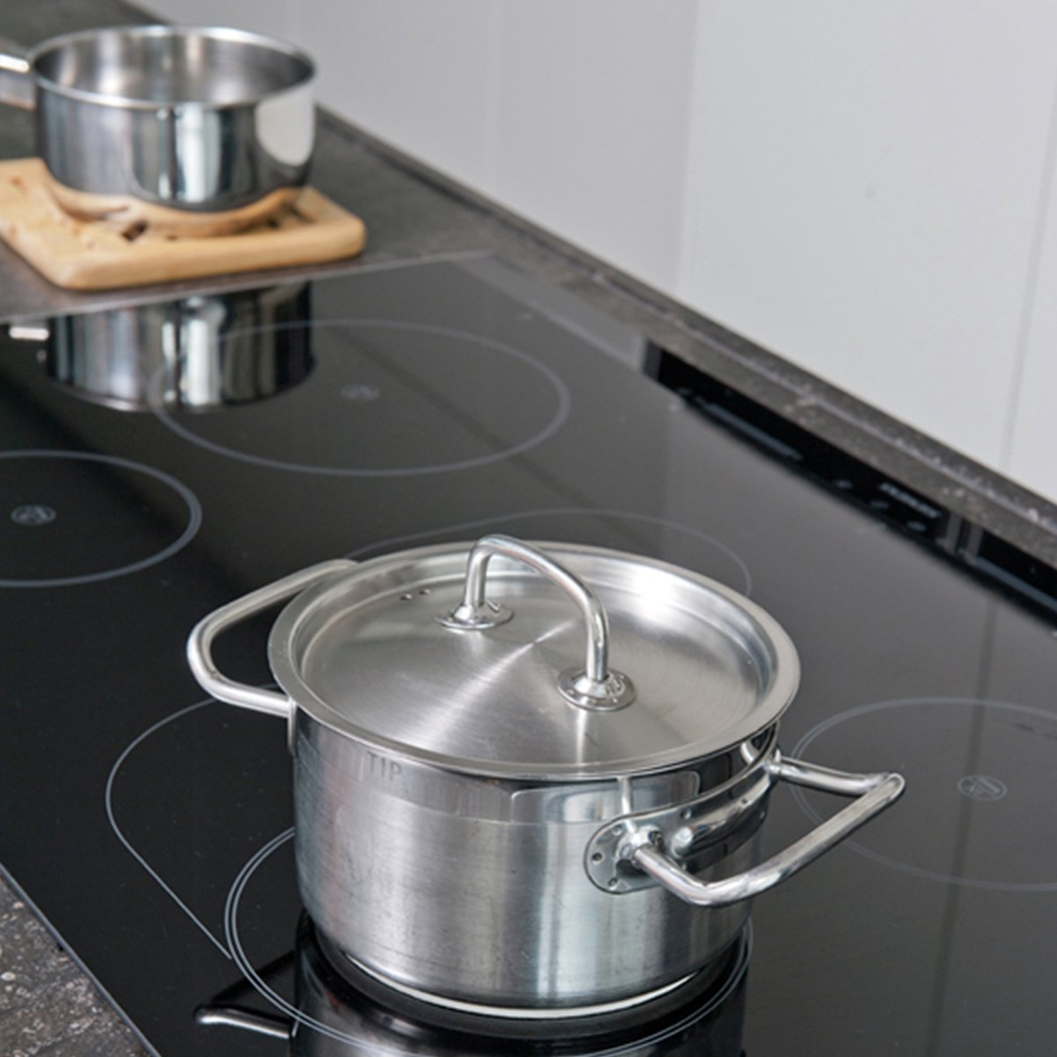 Une casserole en acier inoxydable avec un couvercle est posée sur une plaque de cuisson noire. Une autre casserole sur une planche en bois en arrière-plan.
