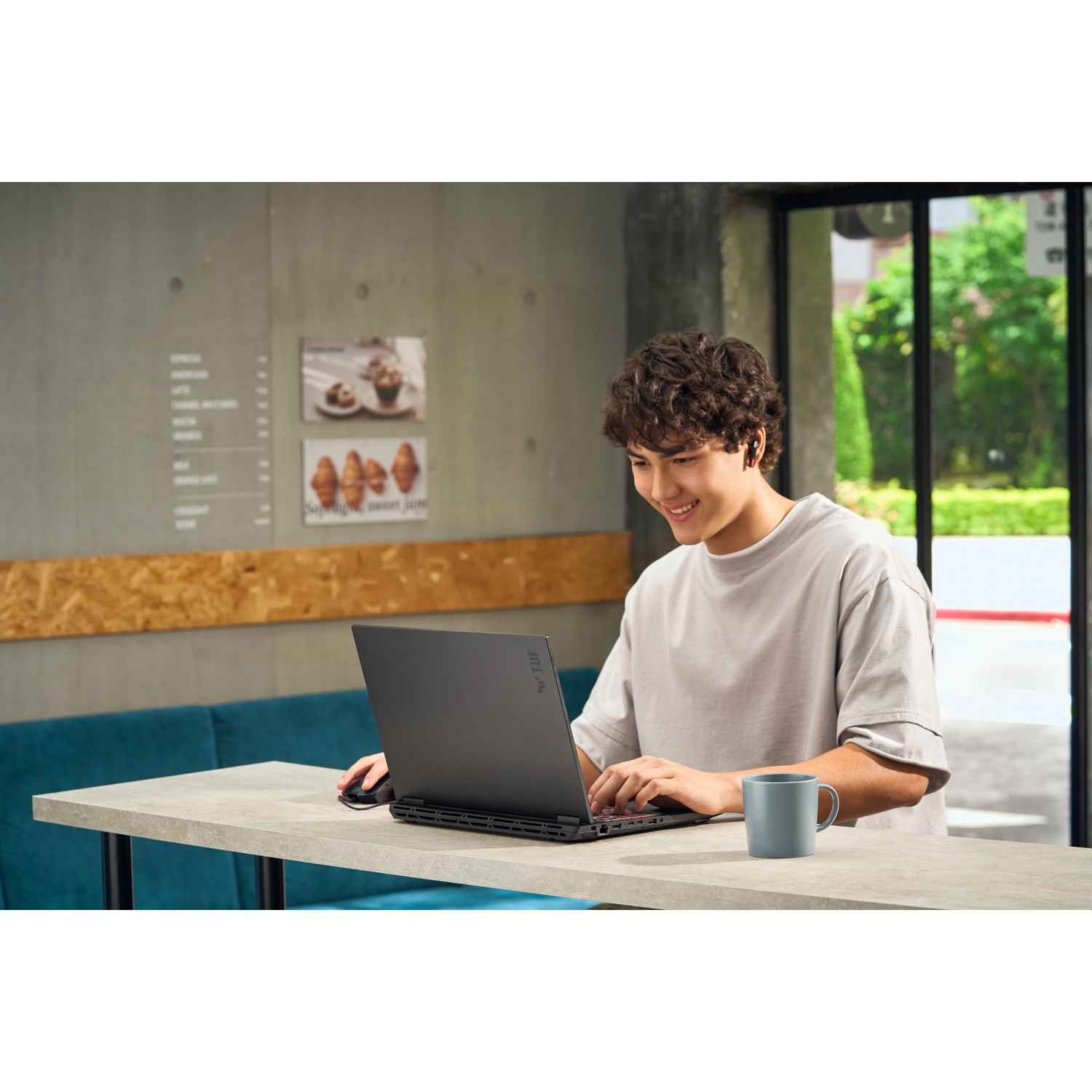 Un jeune homme utilise un ordinateur portable à une table. Il sourit et porte des écouteurs. Une tasse est à côté.