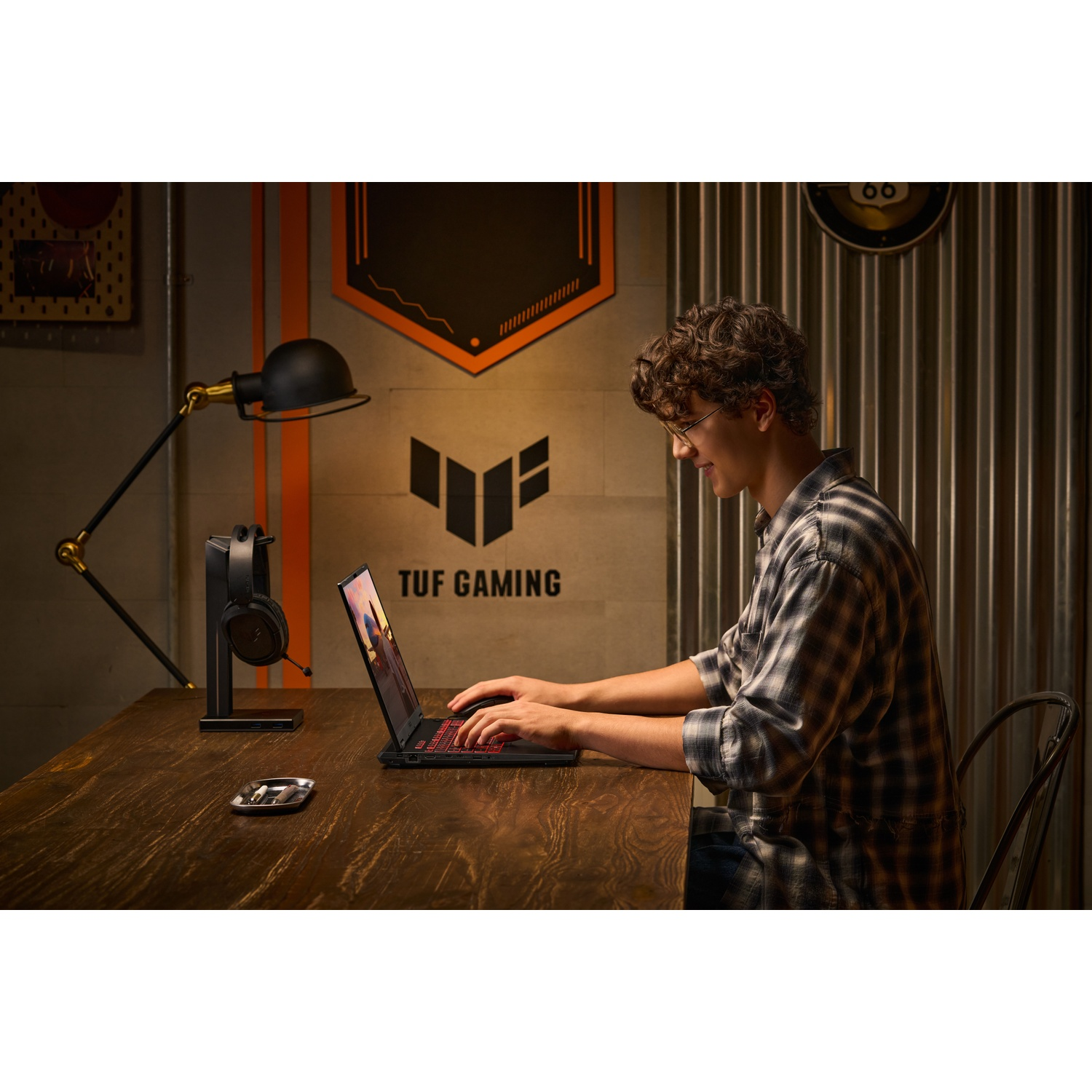 Une personne avec des lunettes utilise un ordinateur portable. L'ordinateur portable a un clavier rouge. Il y a un casque et une lampe sur la table. Le fond est un mur.