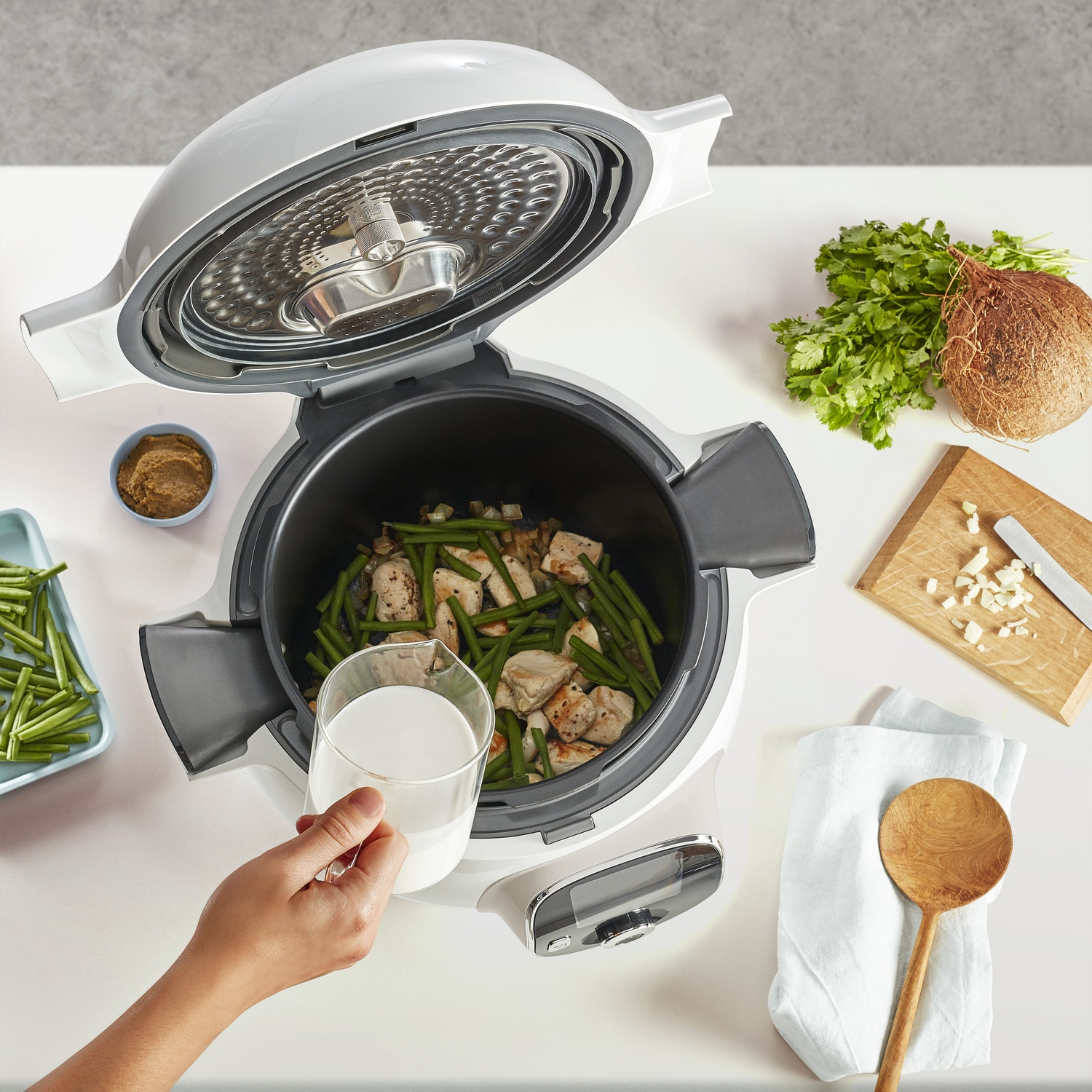 Open multicooker with chicken and green beans. Hand pouring milk. Coconut, cilantro, and spices on the table.