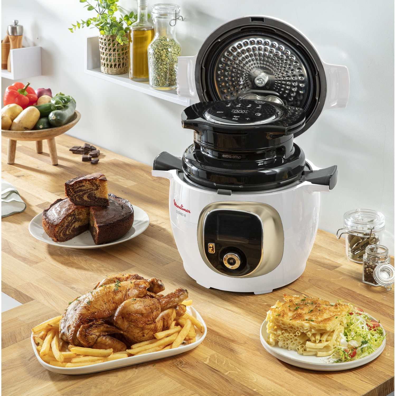 A kitchen scene with a Moulinex Cookeo, food like chicken and cake, and vegetables on a wooden surface.