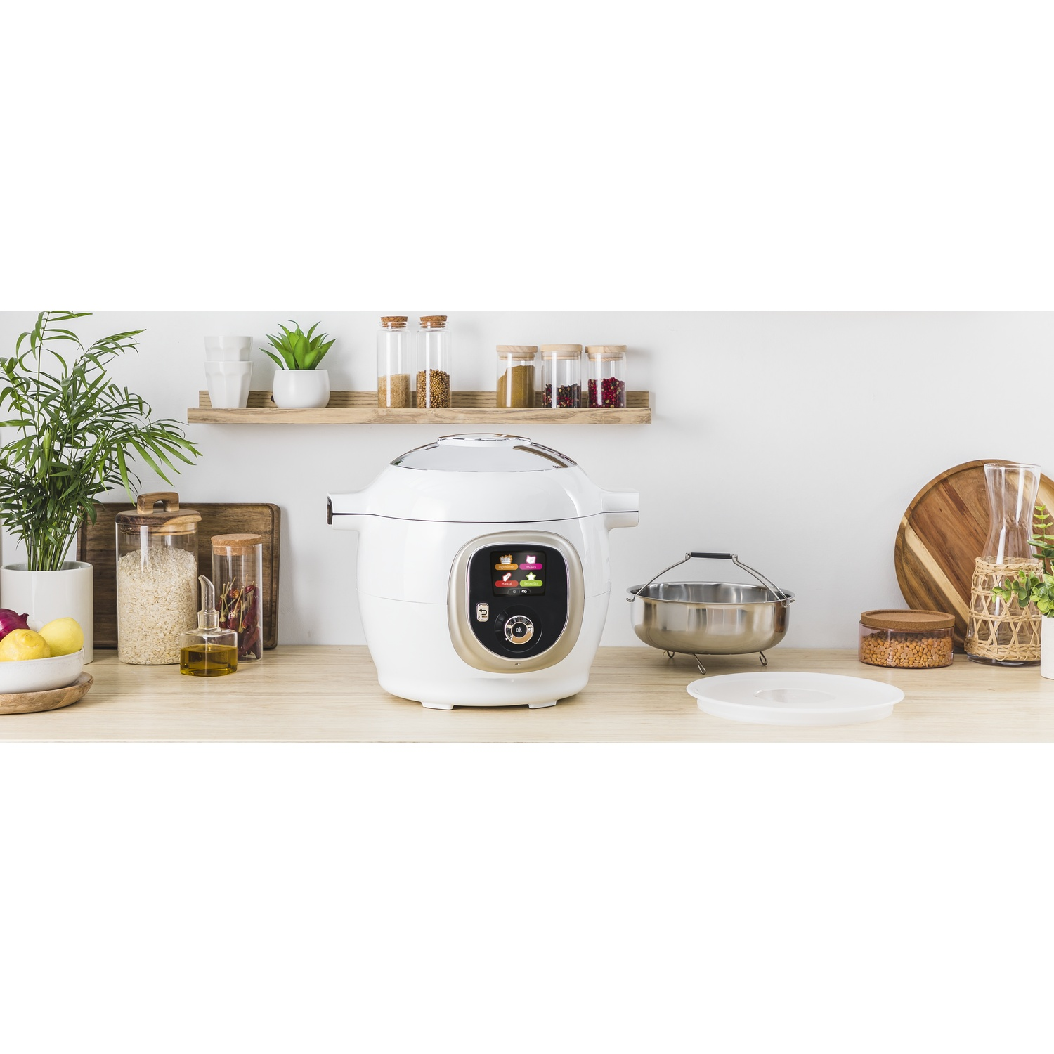 A white kitchen appliance with a digital display sits on a wooden counter. Jars of spices and plants are on a shelf in the background.
