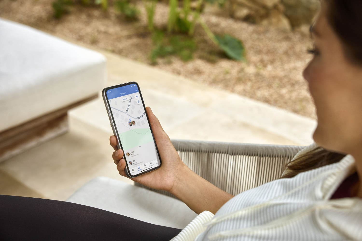 Una mujer sostiene un teléfono que muestra un mapa con ubicaciones. Está al aire libre, sentada, con un fondo borroso de vegetación y un sofá blanco.