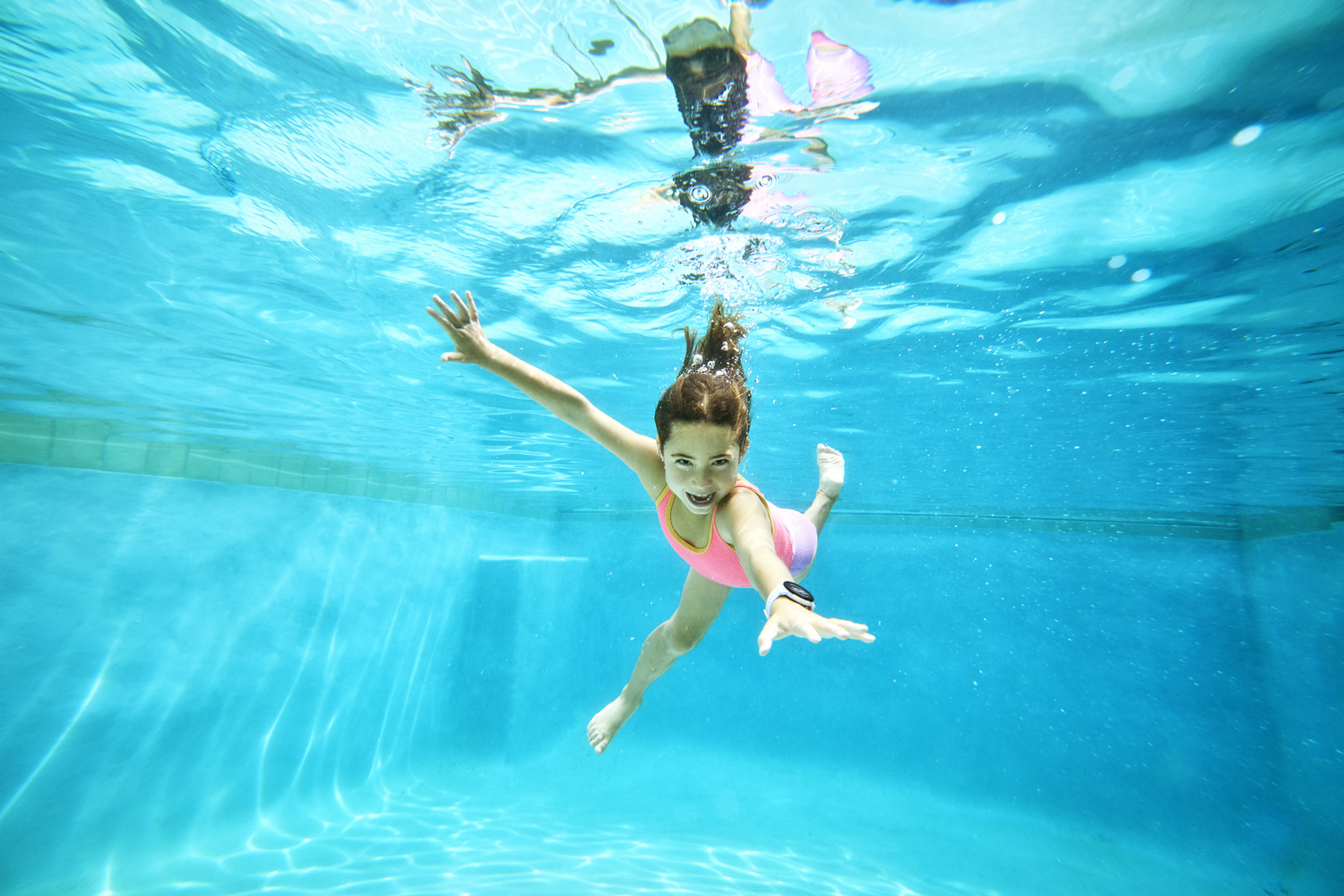 Una niña nada bajo el agua en una piscina, con un traje de baño rosa. Tiene cabello castaño y sonríe, con los brazos extendidos.
