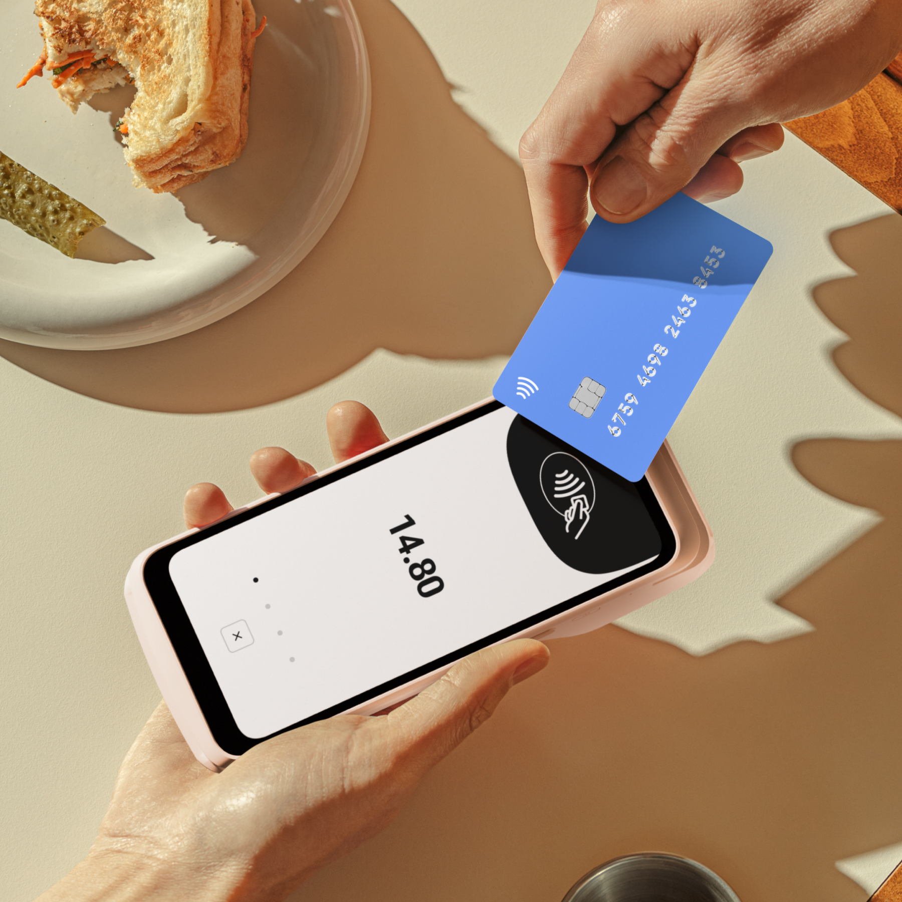 Manos pagando con tarjeta de crédito por teléfono. Un sándwich y un pepinillo están al lado.