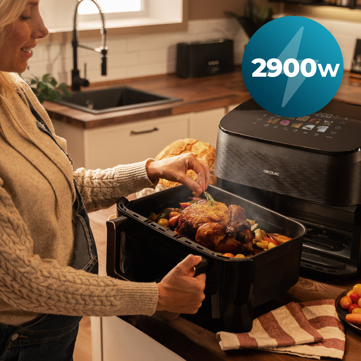 Mujer en la cocina agrega romero al pollo asado en una freidora de aire negra. Se ven verduras. Un símbolo de 2900w está en el fondo.