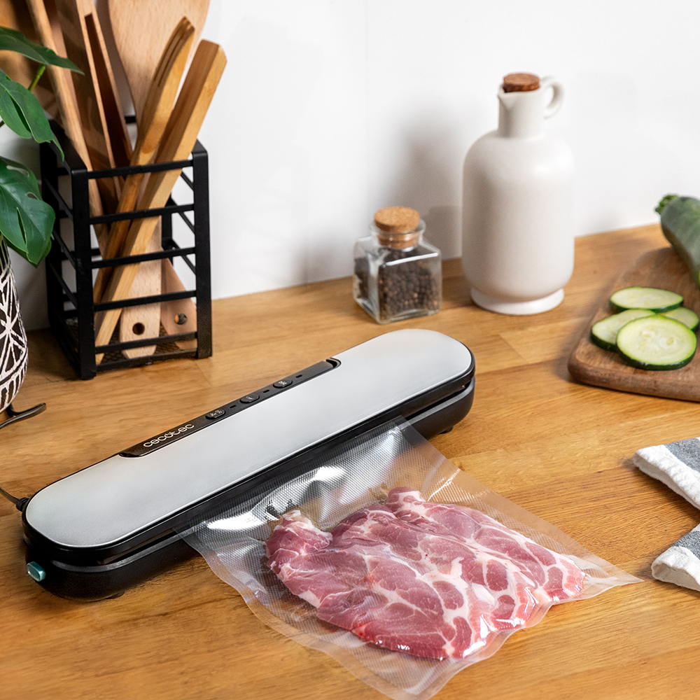 Una envasadora al vacío con carne dentro de una bolsa de plástico sobre una mesa de madera. Otros objetos incluyen un calabacín, un tarro y utensilios.