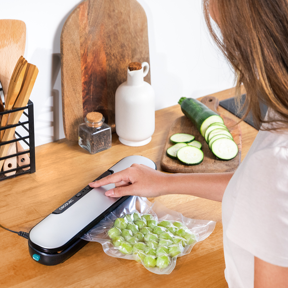 Una mujer usa una envasadora al vacío en una bolsa de comida verde. Calabacín y otros utensilios de cocina están al fondo.