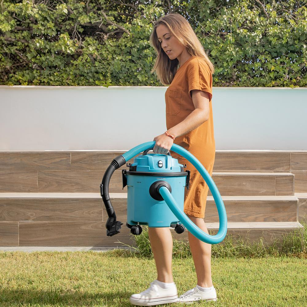 Una mujer con vestido naranja lleva una aspiradora azul sobre césped. En el fondo, escaleras y follaje verde.