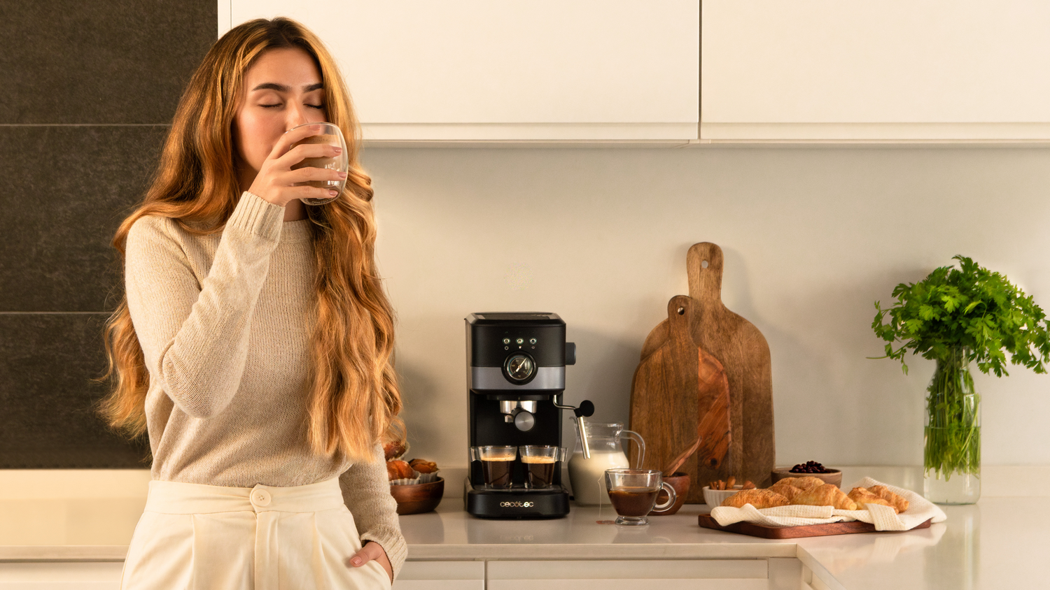Mujer bebiendo de un vaso. Cafetera, cruasanes y hierbas en la encimera de una cocina.