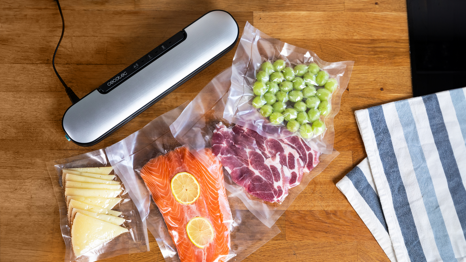 Alimentos sellados: uvas, carne, salmón con limones, queso. Selladora al vacío sobre mesa de madera, tela a rayas.