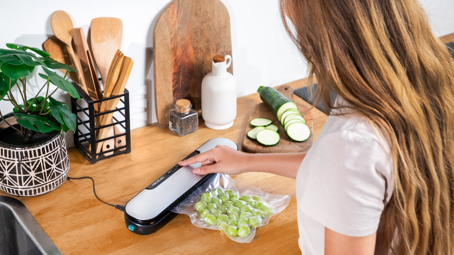 Una mujer usa una envasadora al vacío en una bolsa de uvas. Calabacín y utensilios de cocina en la encimera de madera.