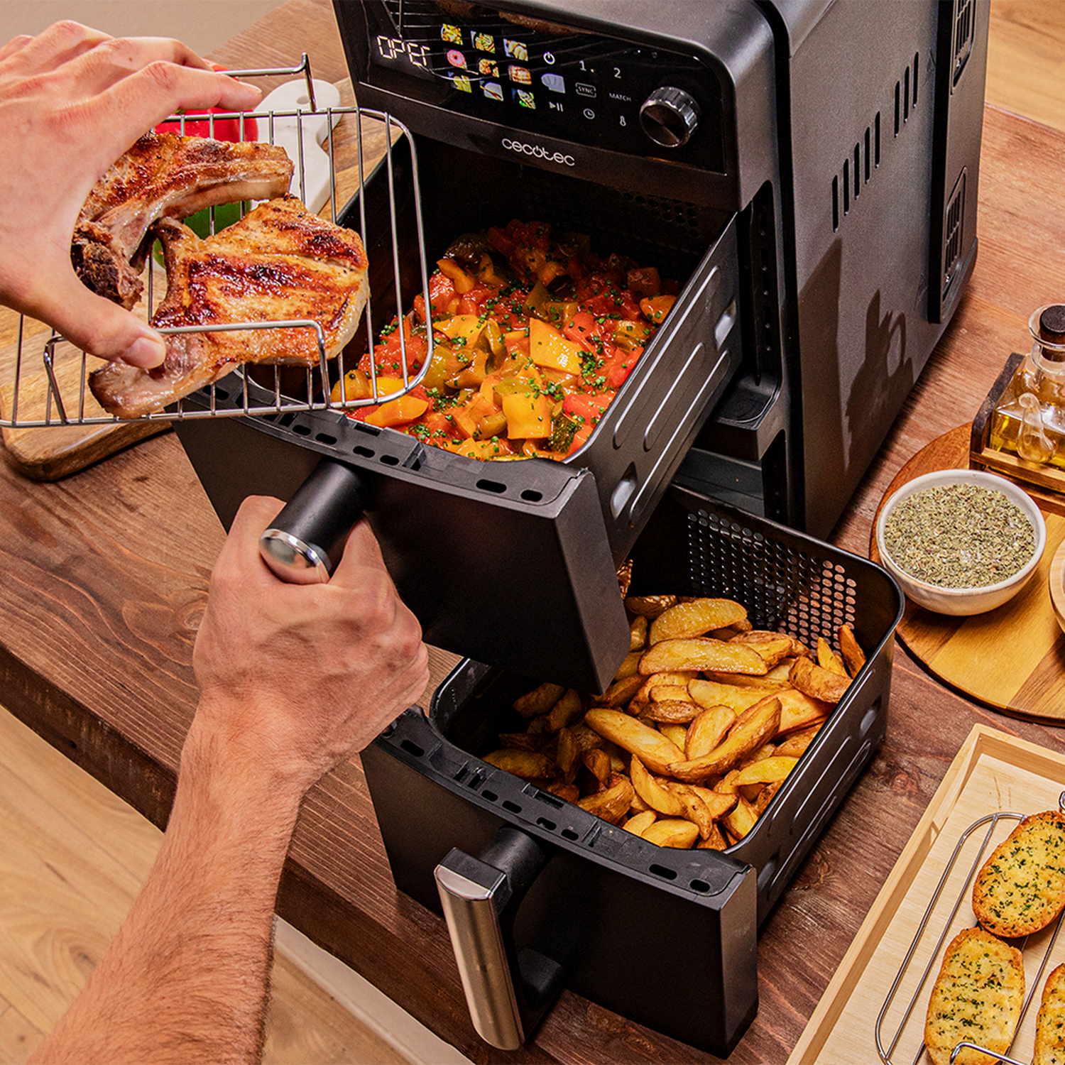 Una freidora de aire con comida dentro, una mano sosteniendo una rejilla de costillas cocinadas. También se ven papas fritas y verduras.