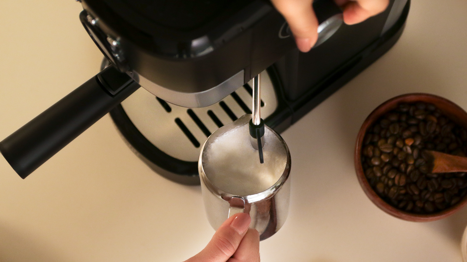 Una cafetera espumando leche en una jarra de metal. Granos de café en un cuenco de madera cercanos.