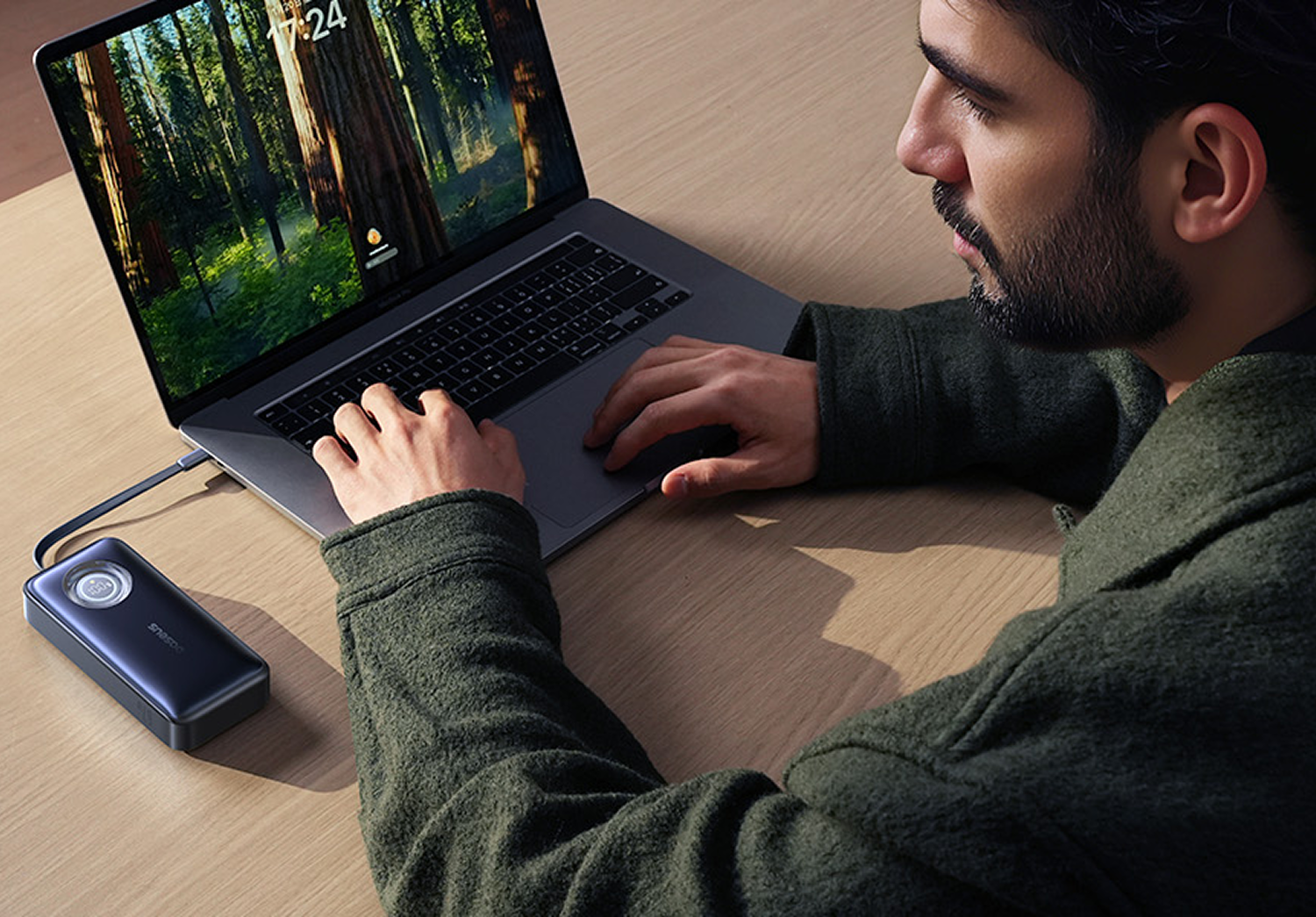 Un hombre usando una laptop, conectado a un banco de energía. La laptop está sobre una mesa de madera. La pantalla muestra un bosque.