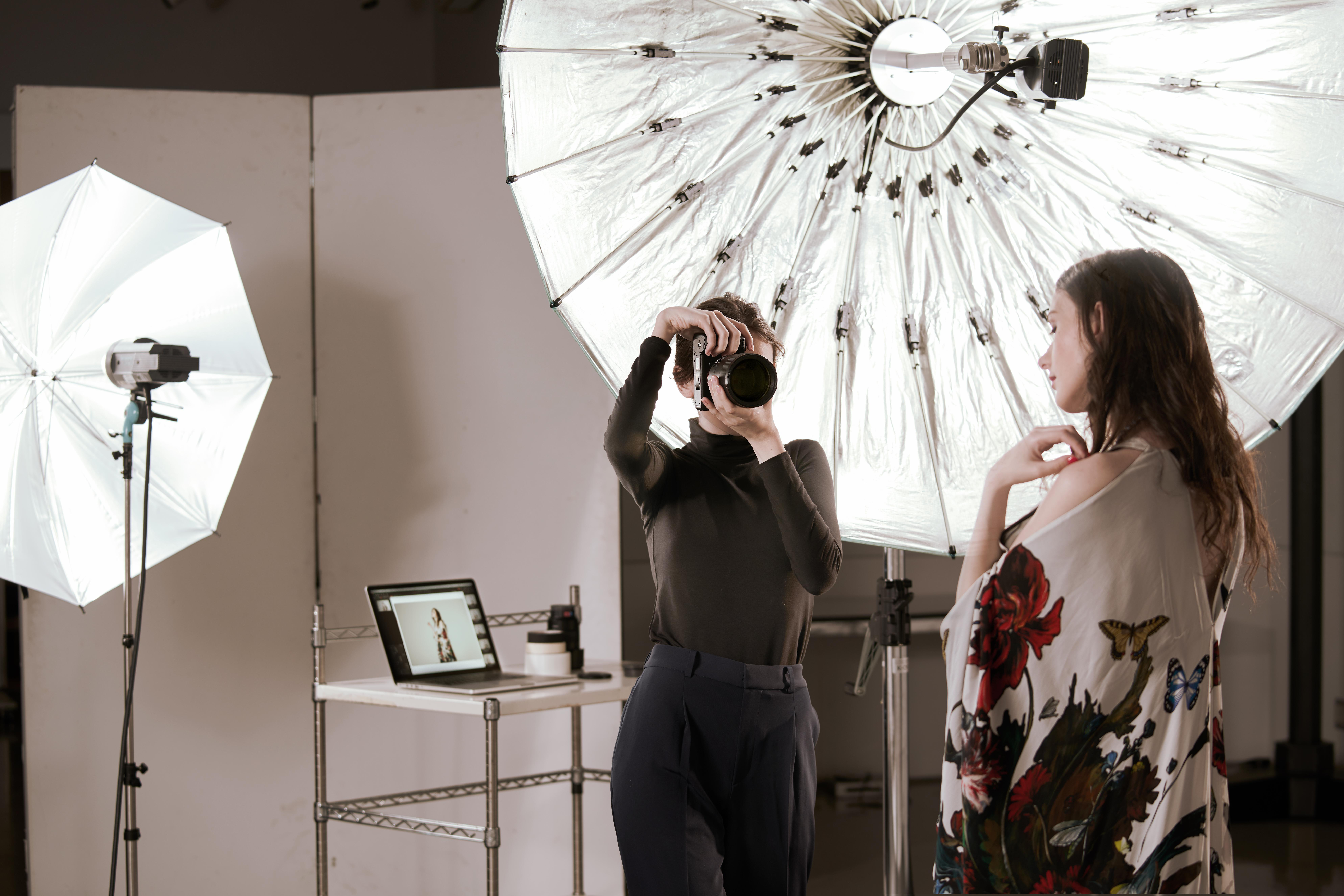 Ein Fotograf fotografiert ein Model im Studio. Das Model trägt ein Oberteil mit Blumenmuster. Beleuchtung sichtbar.