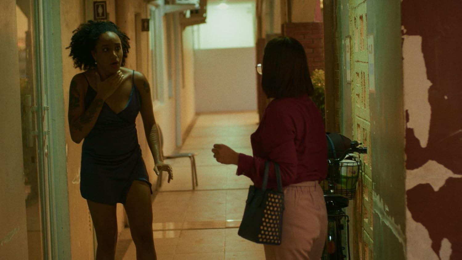 Zwei Frauen in einem Flur. Eine in einem blauen Kleid sieht geschockt aus. Die andere in rosa Hose hält eine Handtasche. Im Hintergrund ein Fahrrad.