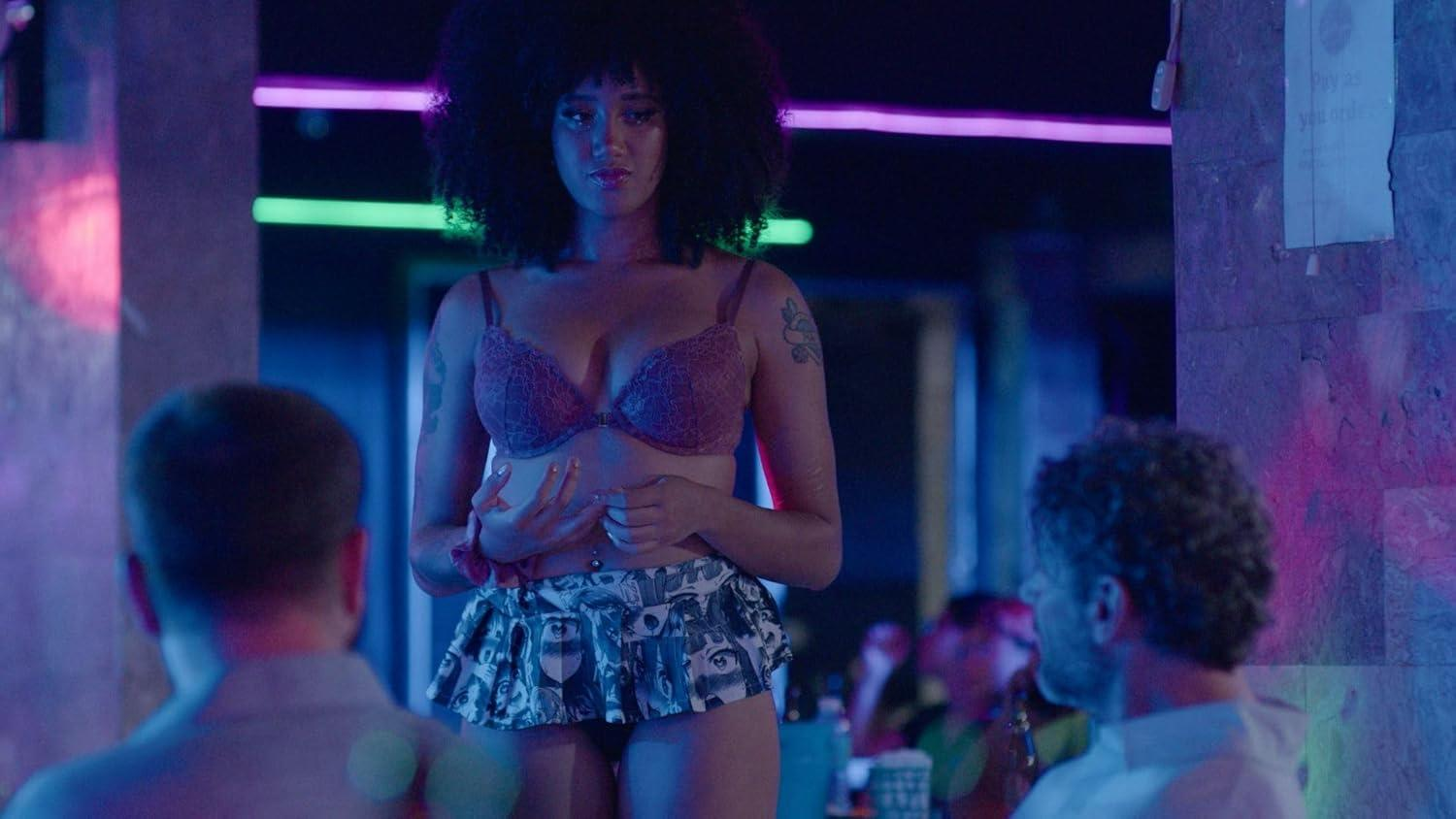 Eine Frau in Dessous steht in einem Club. Zwei Männer sitzen im Vordergrund. Lila und grüne Neonlichter sind im Hintergrund sichtbar.