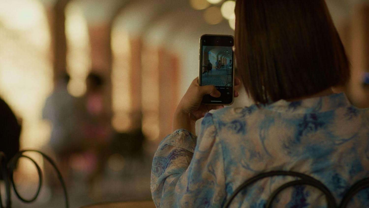 Eine Person hält ein Telefon und macht ein Foto. Das Telefon zeigt eine Innenszene. Die Person trägt ein blaues Blumenhemd.