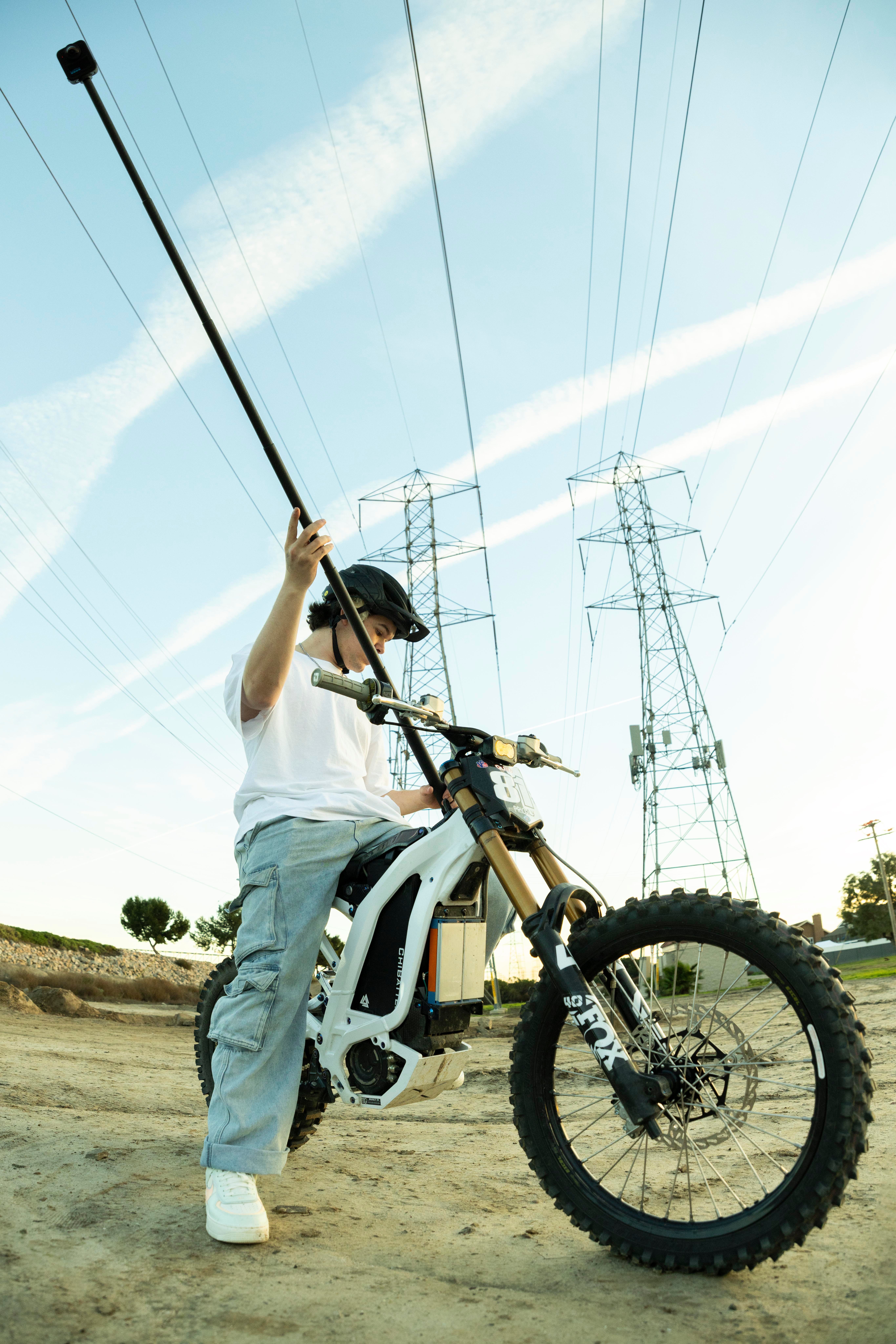 Eine Person auf einem Elektromotorrad hält eine lange Stange. Stromleitungen und -masten sind im Hintergrund.