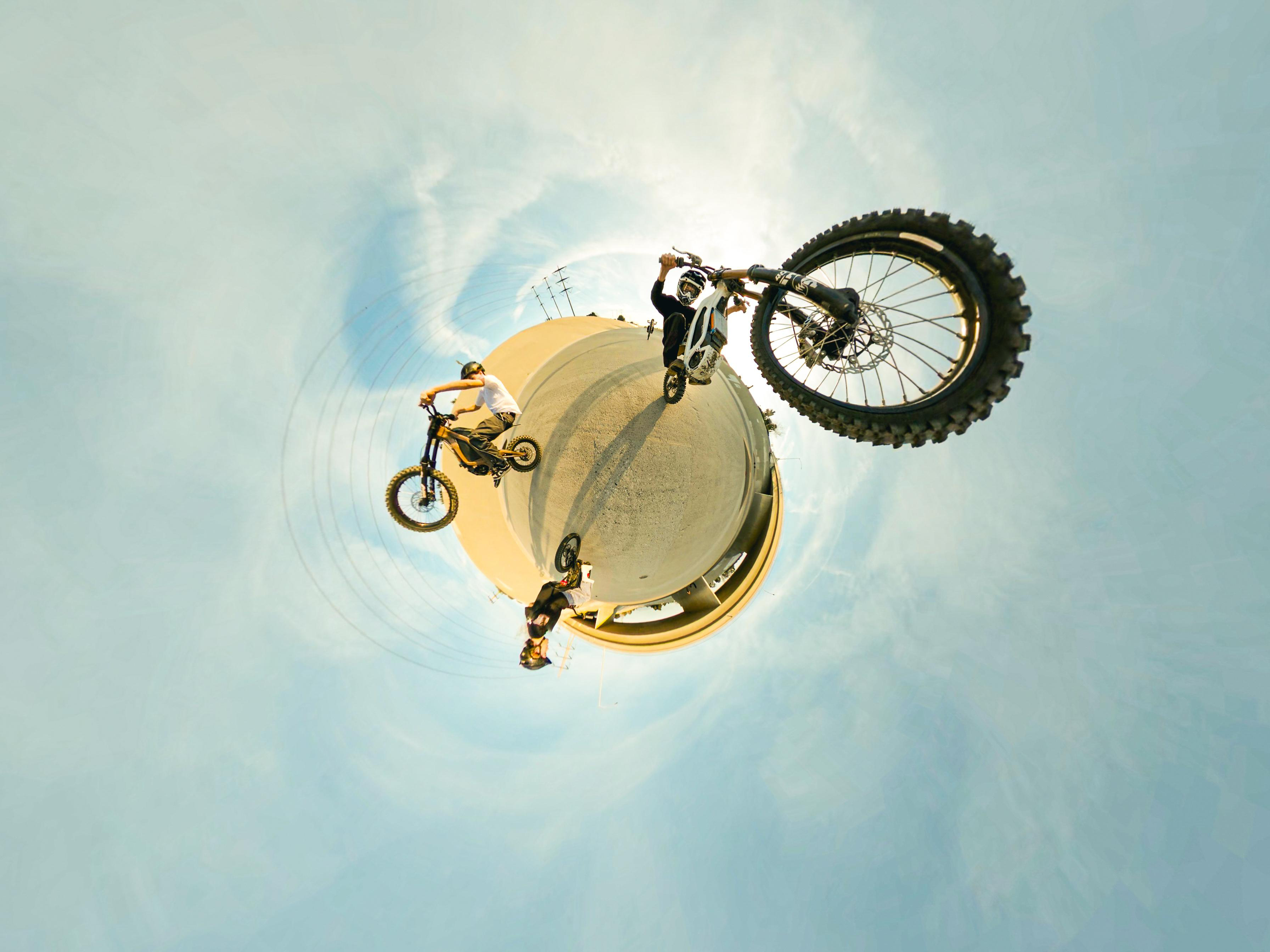 Drei Personen auf Motorrädern, die Stunts auf einer gekrümmten Oberfläche vor einem bewölkten Himmel machen.