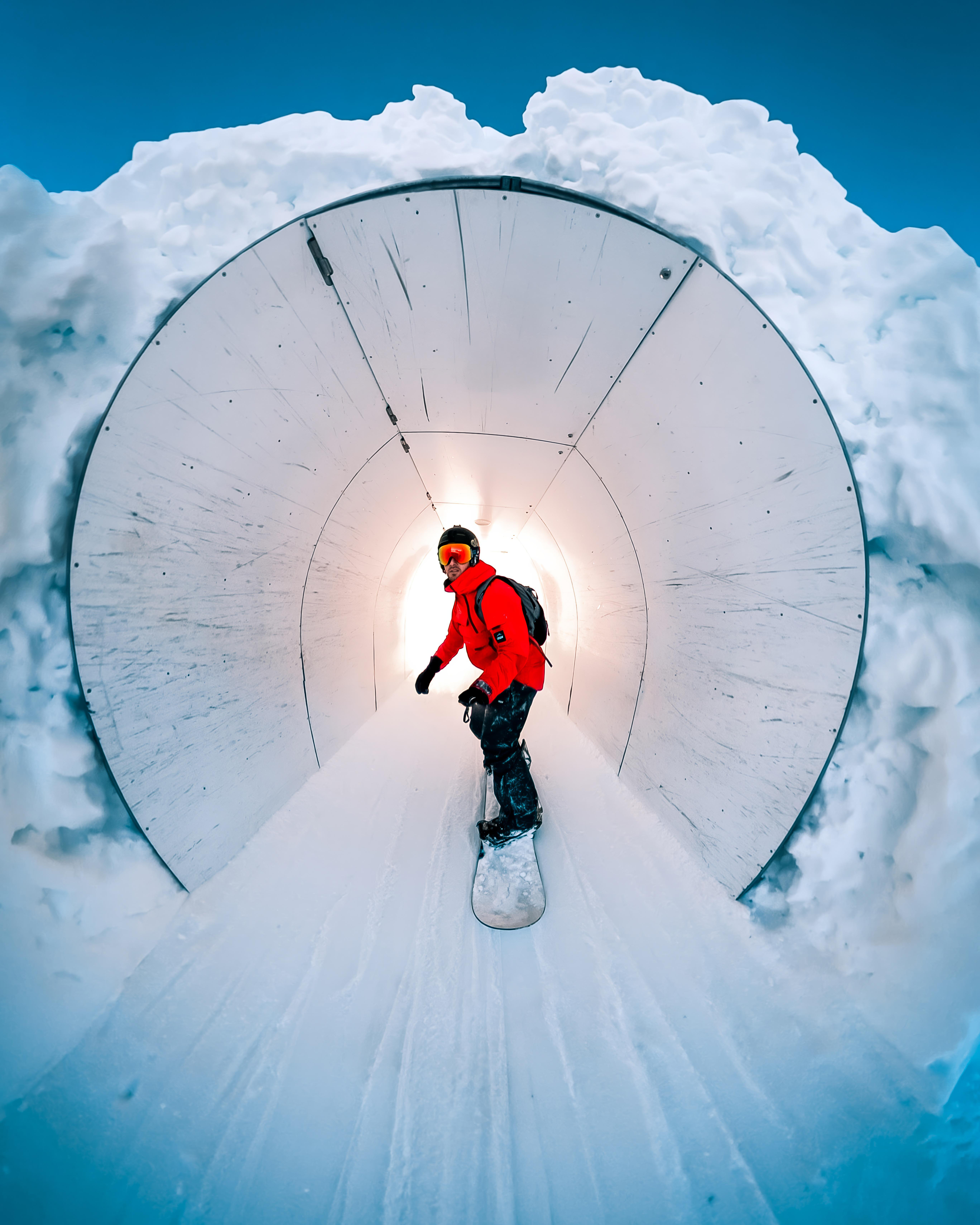 Snowboarder in roter Jacke in einem Schneetunnel, umgeben von Schnee und blauem Himmel.