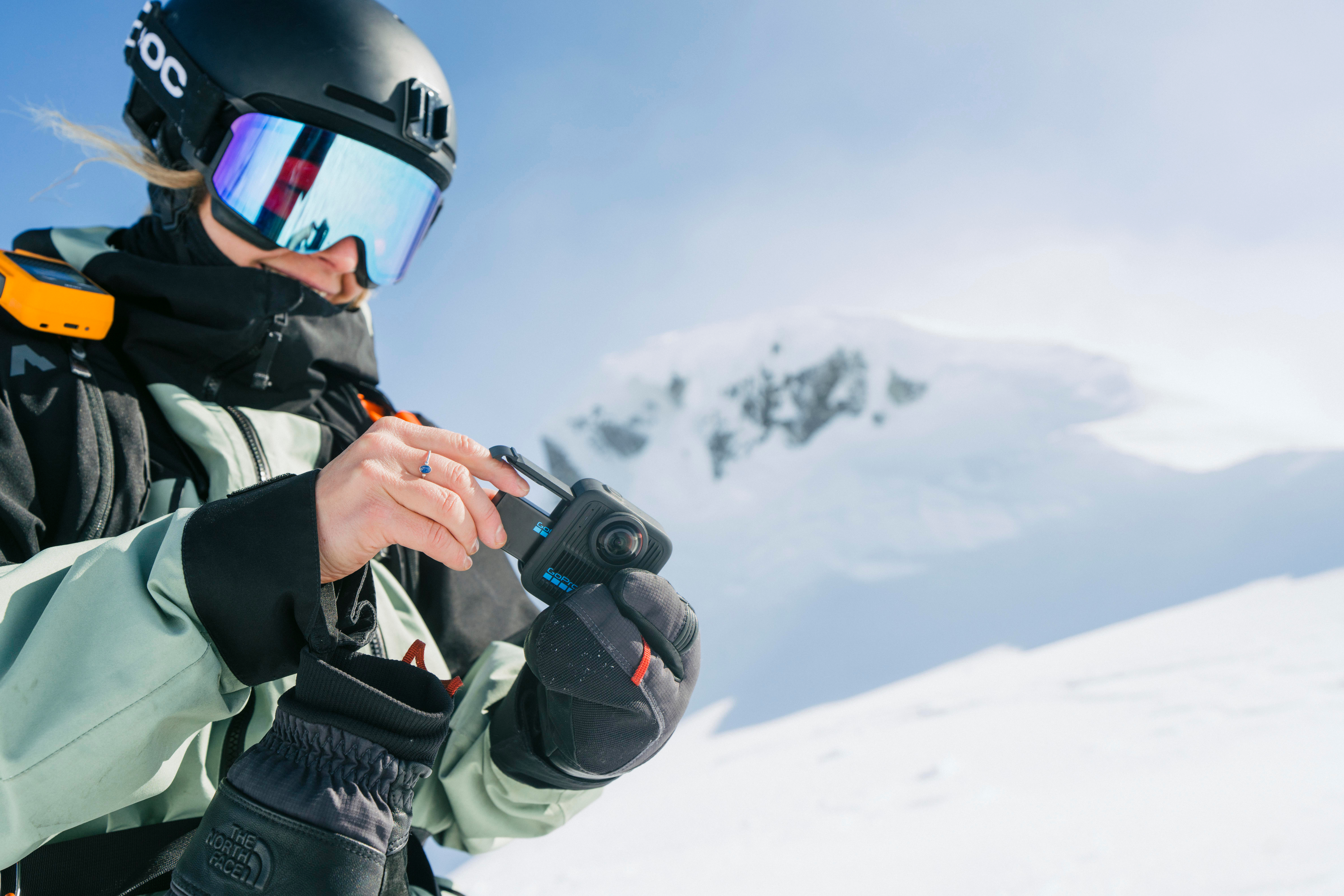 Skifahrer hält GoPro-Kamera, trägt Brille und Helm, mit schneebedecktem Berg und blauem Himmel.