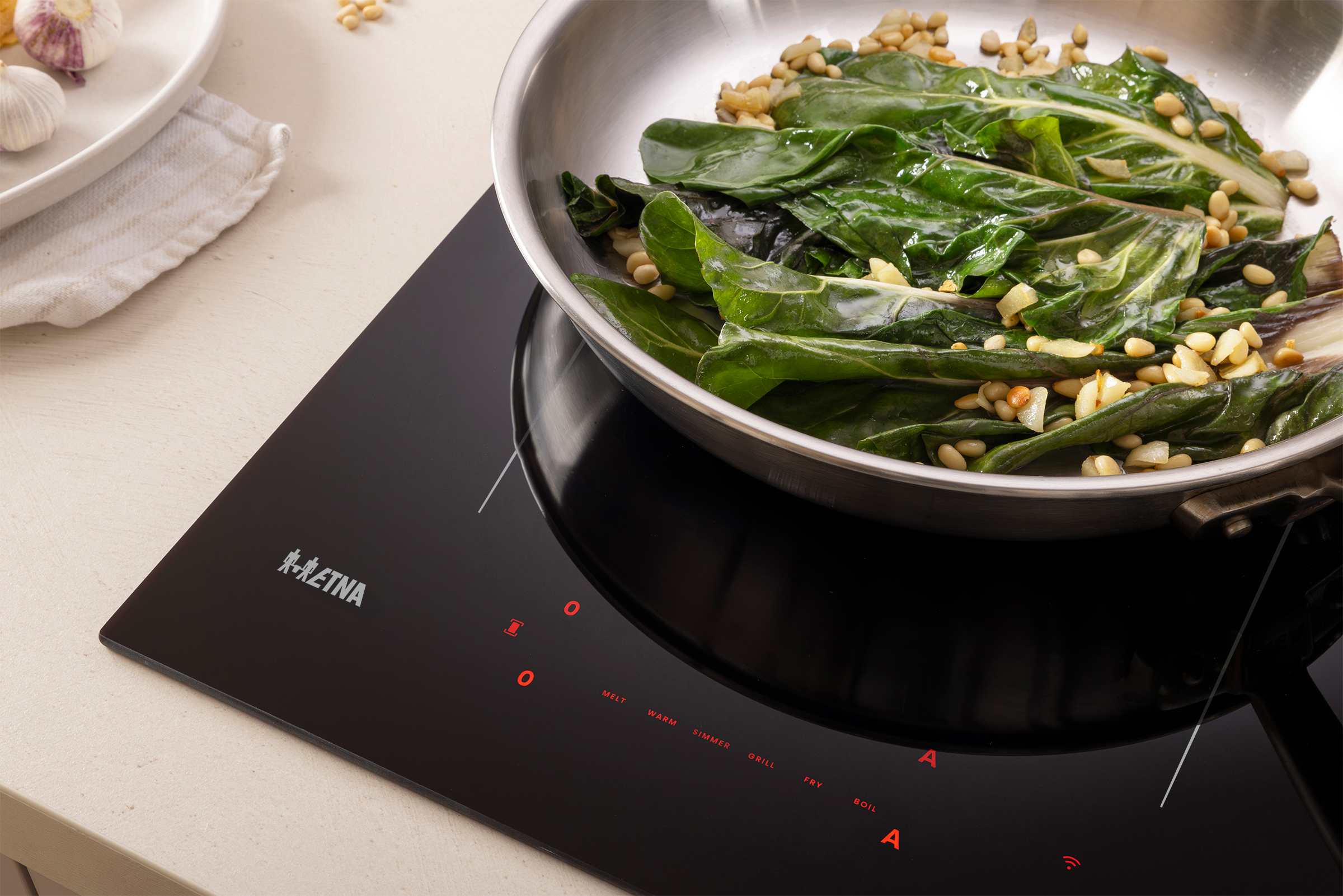 A pan with cooked greens and nuts sits on a black stovetop. Garlic and a plate are on the side.