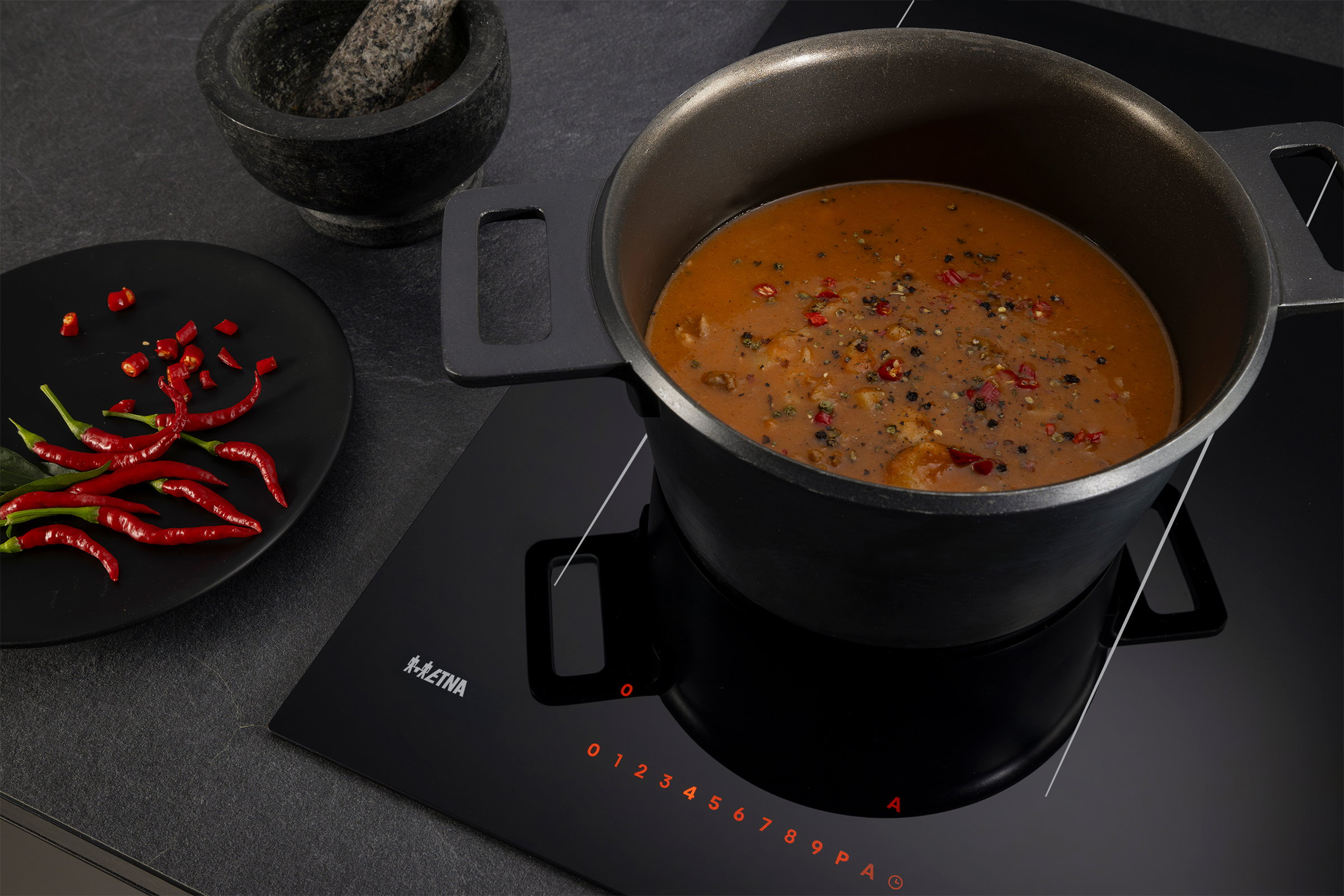 A pot of soup on a stove, with red peppers on a plate. A mortar and pestle are on the side.