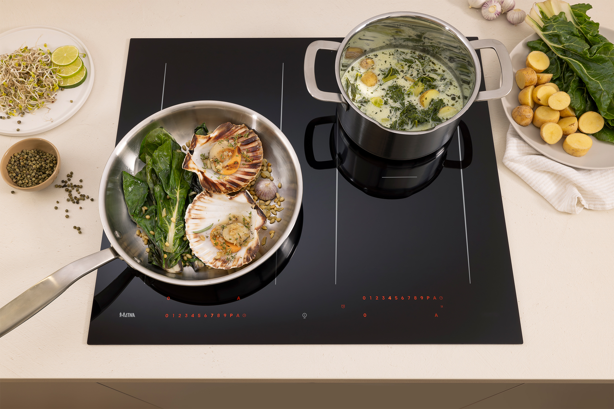 A black hob with cooking ingredients. A pan with scallops, a pot of soup, plates of food, and garlic.