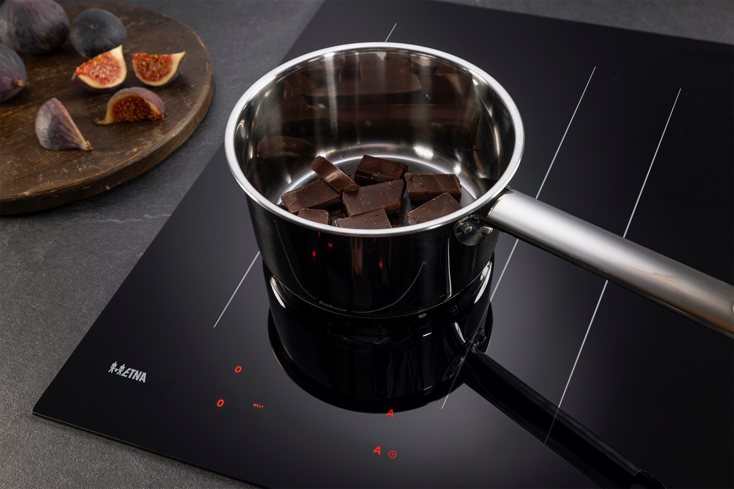 A stainless steel pot with chocolate chunks sits on a black stovetop. Figs are on a wooden board.
