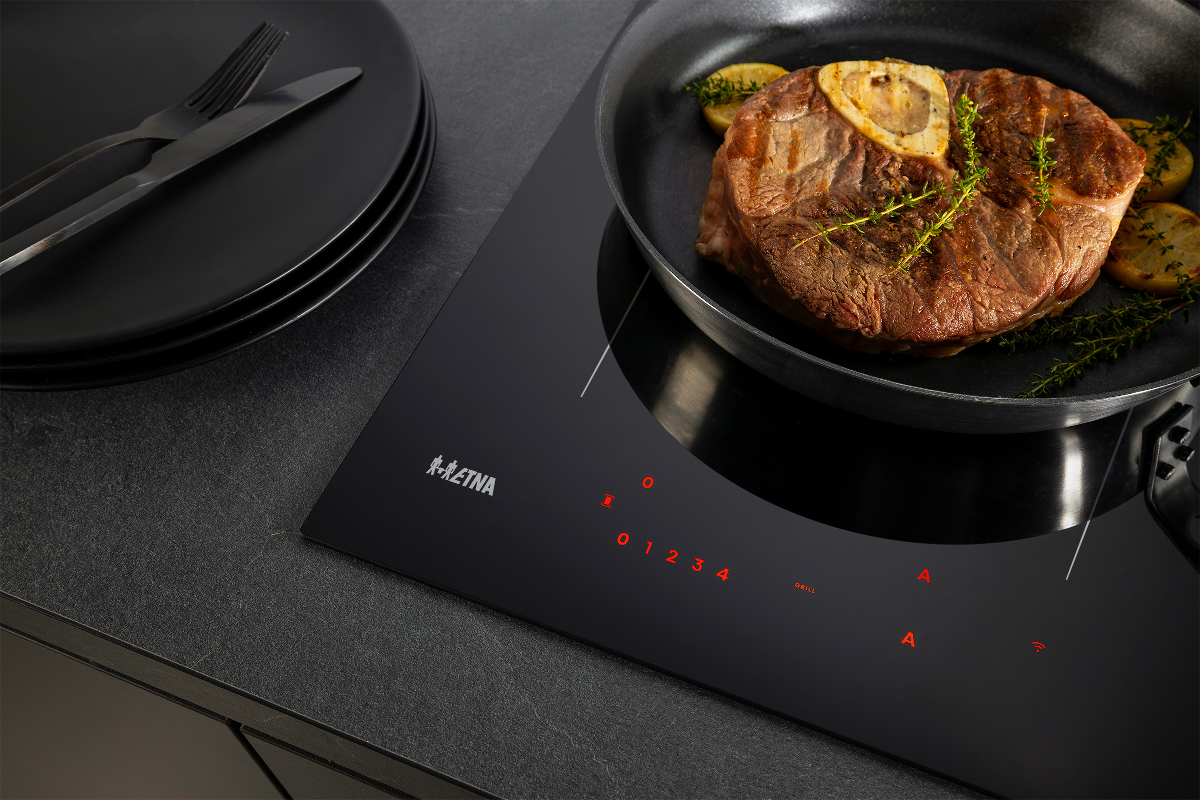 A steak in a pan on a black stove. Black plates with cutlery are on the left. The stove has red controls.