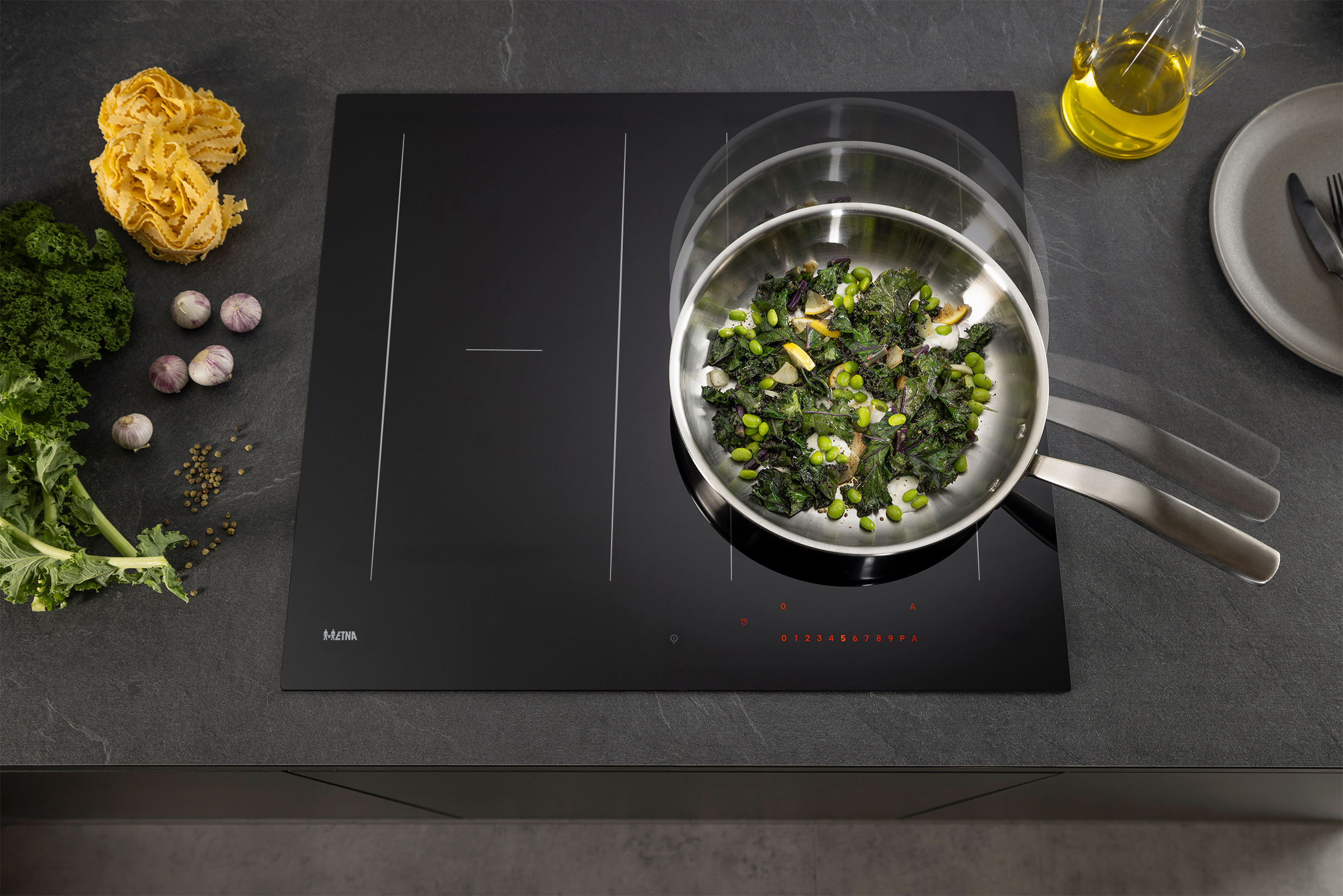 A pan with food on a black stovetop, surrounded by ingredients like pasta, garlic, and oil, on a dark countertop.