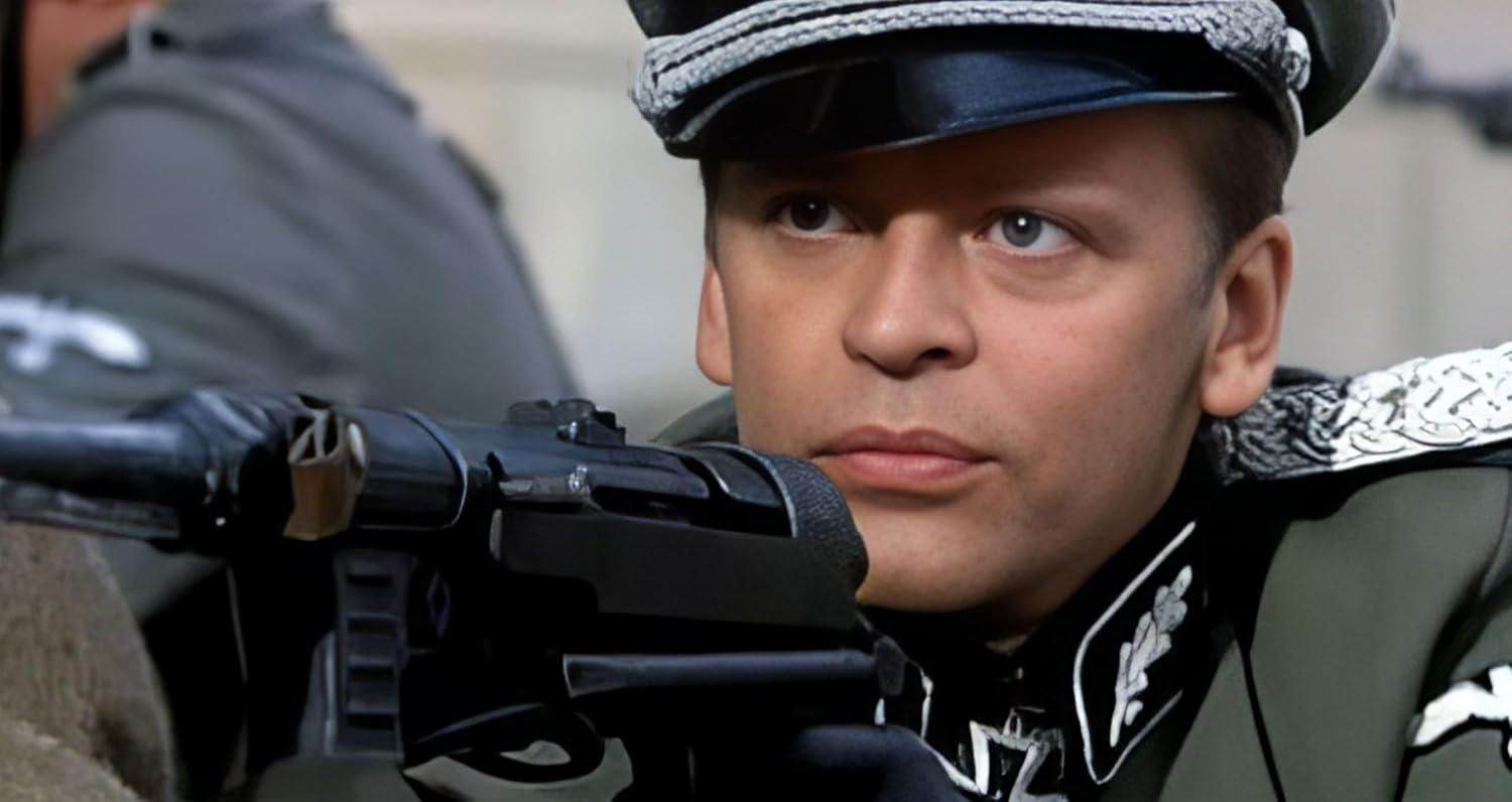 Ein Mann in Uniform hält eine Maschinenpistole und trägt eine schwarze Mütze. Der Mann hat einen ernsten Gesichtsausdruck und schaut nach vorne.