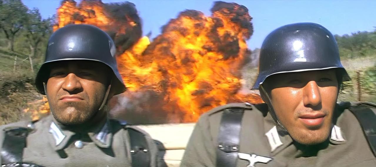 Zwei Soldaten in Helmen und Uniformen. Im Hintergrund eine große Explosion. Blauer Himmel und Bäume sind sichtbar.
