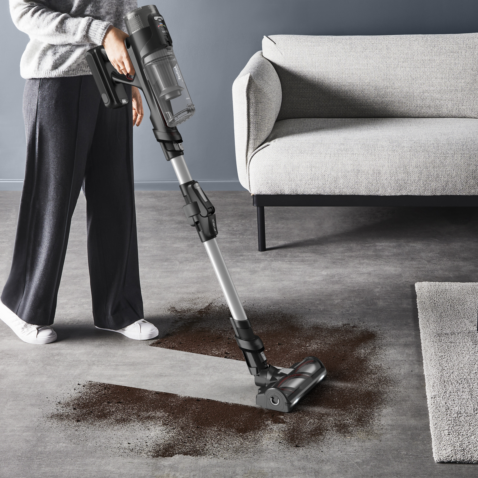 Osoba odkurza rozlaną ziemię odkurzaczem bezprzewodowym. W tle kanapa i dywan.