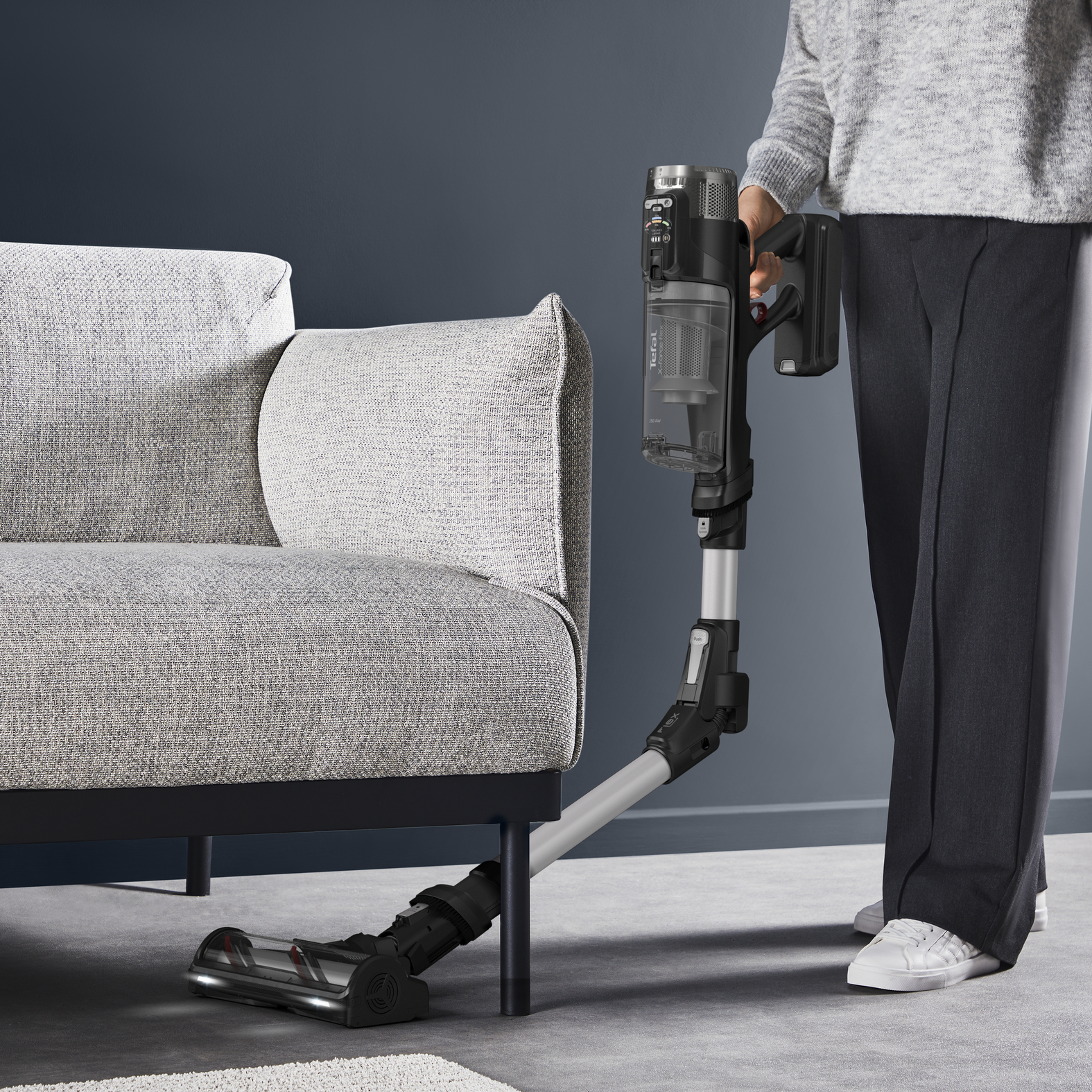 Osoba odkurza szary dywan czarnym odkurzaczem bezprzewodowym w pobliżu szarej sofy.