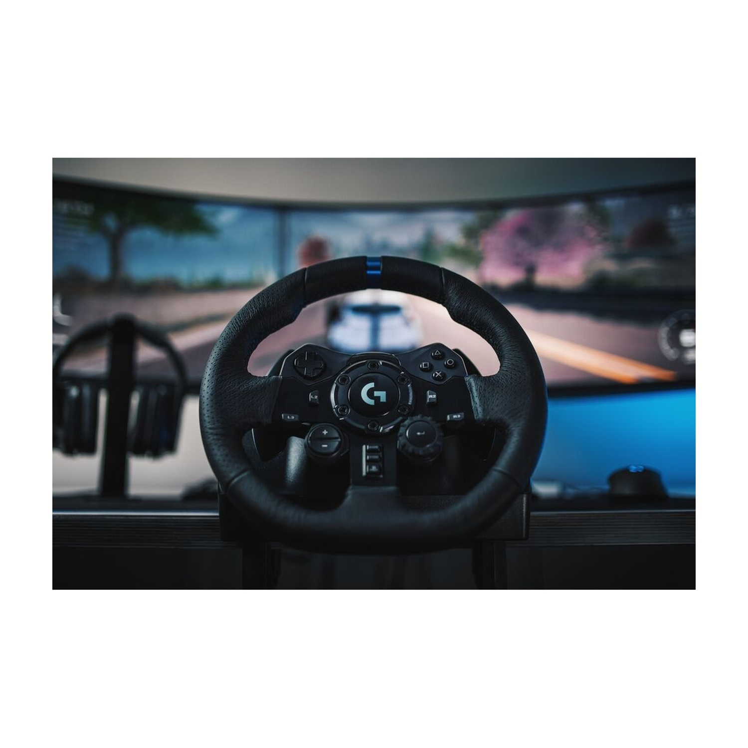 Un volante da corsa Logitech nero con pulsanti, uno schermo con un gioco di corse automobilistiche e cuffie sullo sfondo.