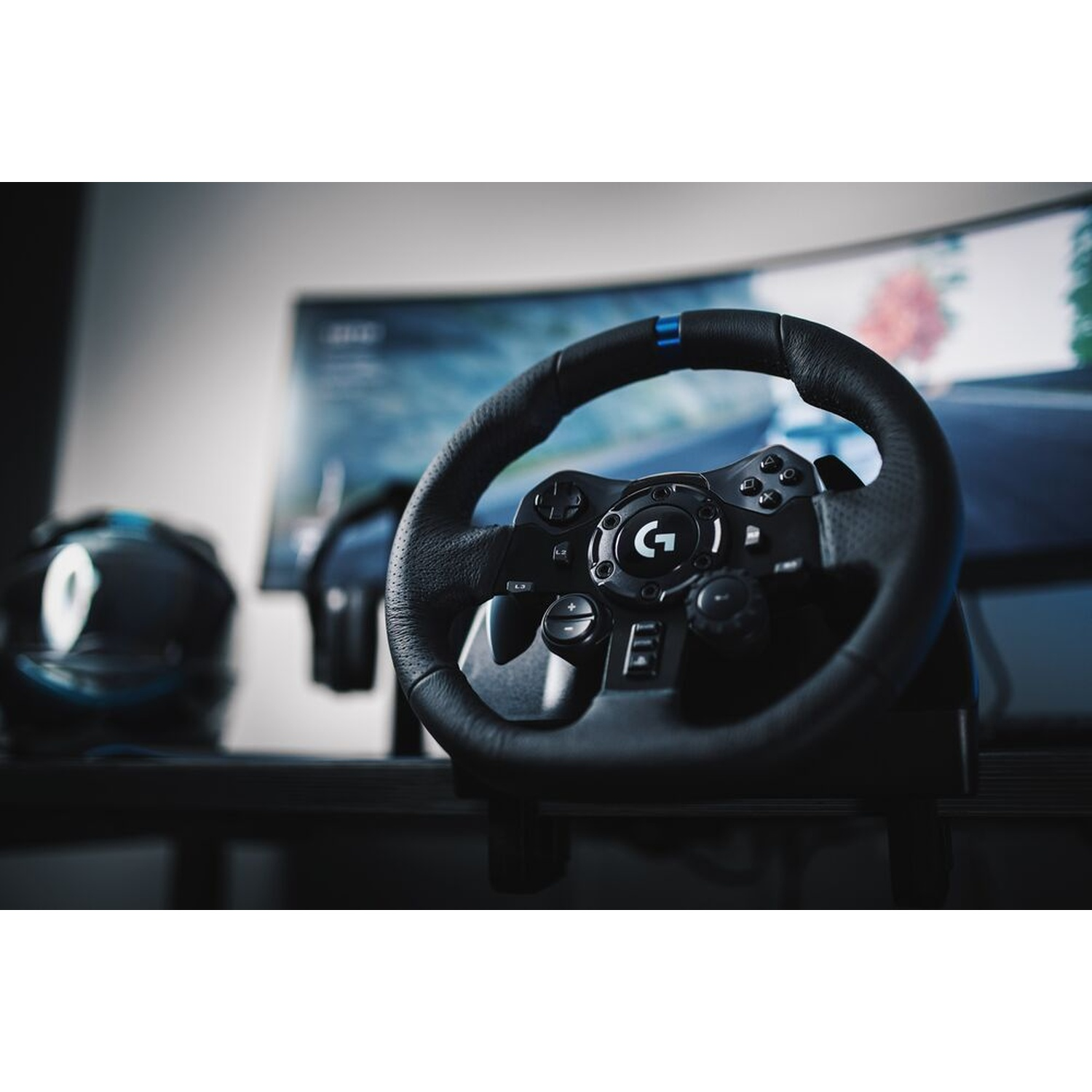 Un volante da gioco Logitech nero è a fuoco. Sullo sfondo un monitor con un gioco di corse e altri accessori.