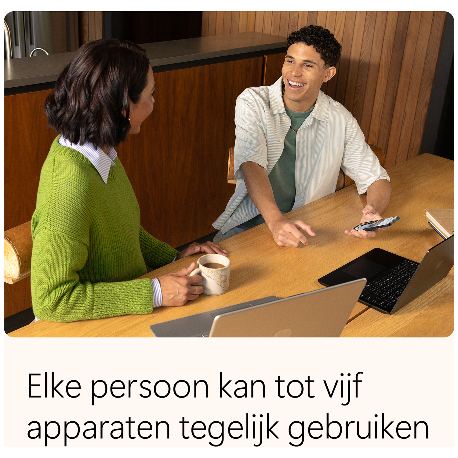 Two people at a wooden table smiling. A woman holds a mug. Laptops and a phone are on the table.