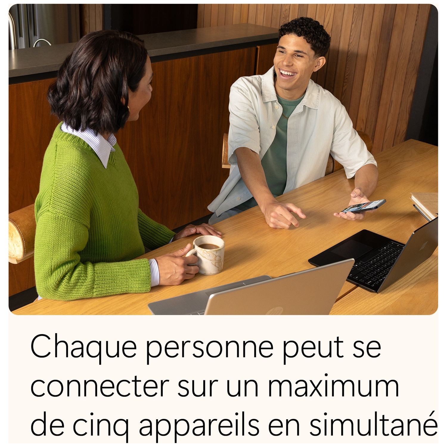Deux personnes à une table en bois avec des ordinateurs portables et un téléphone. L'un tient une tasse, l'autre sourit. L'intérieur.