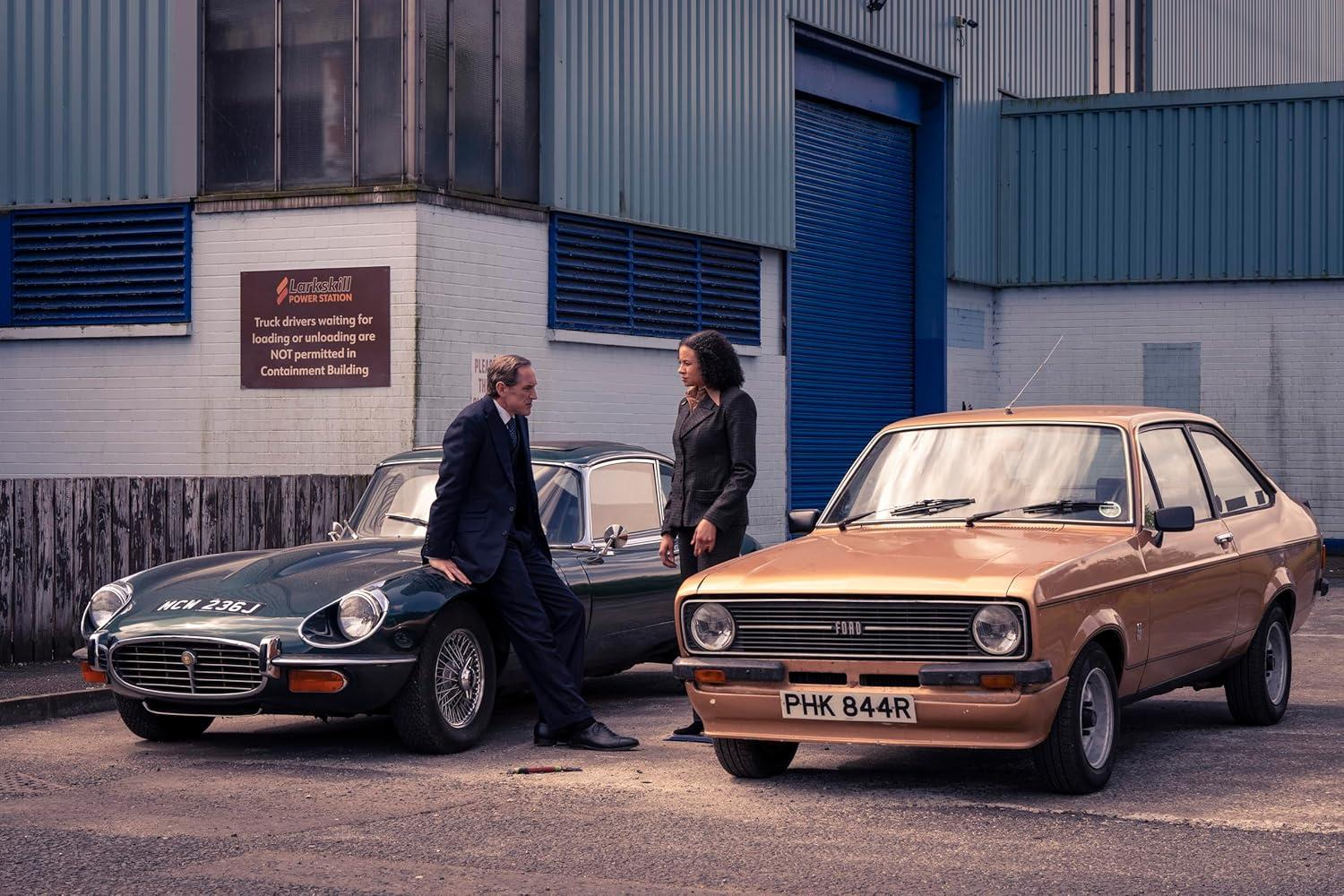 Ein Mann sitzt auf einem grünen Jaguar neben einer Frau und einem braunen Ford. Im Hintergrund ist ein Kraftwerk zu sehen.