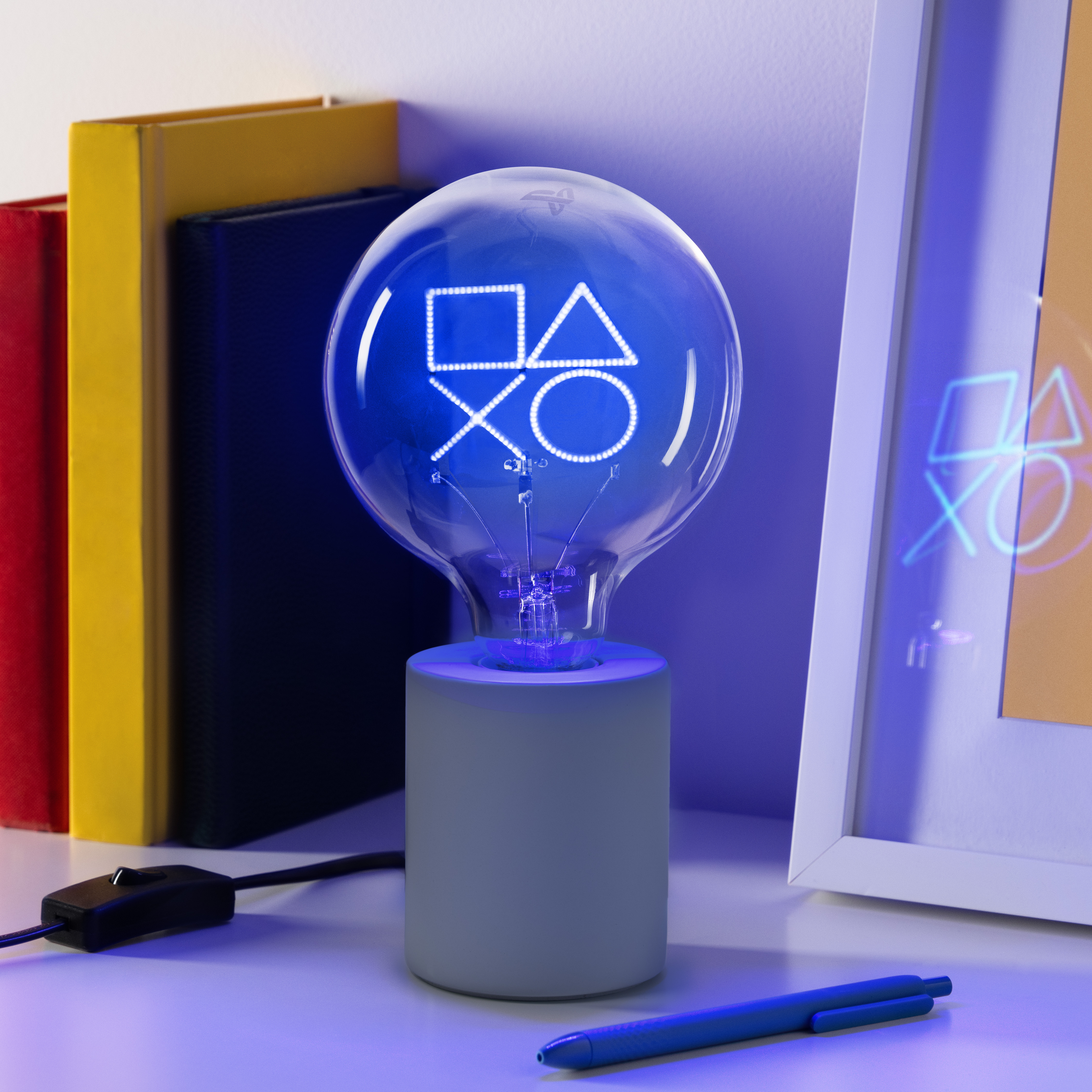 Eine PlayStation-Logo-Glühbirne steht auf einem Tisch mit Büchern, einem Rahmen und einem Stift. Die Glühbirne leuchtet blau.
