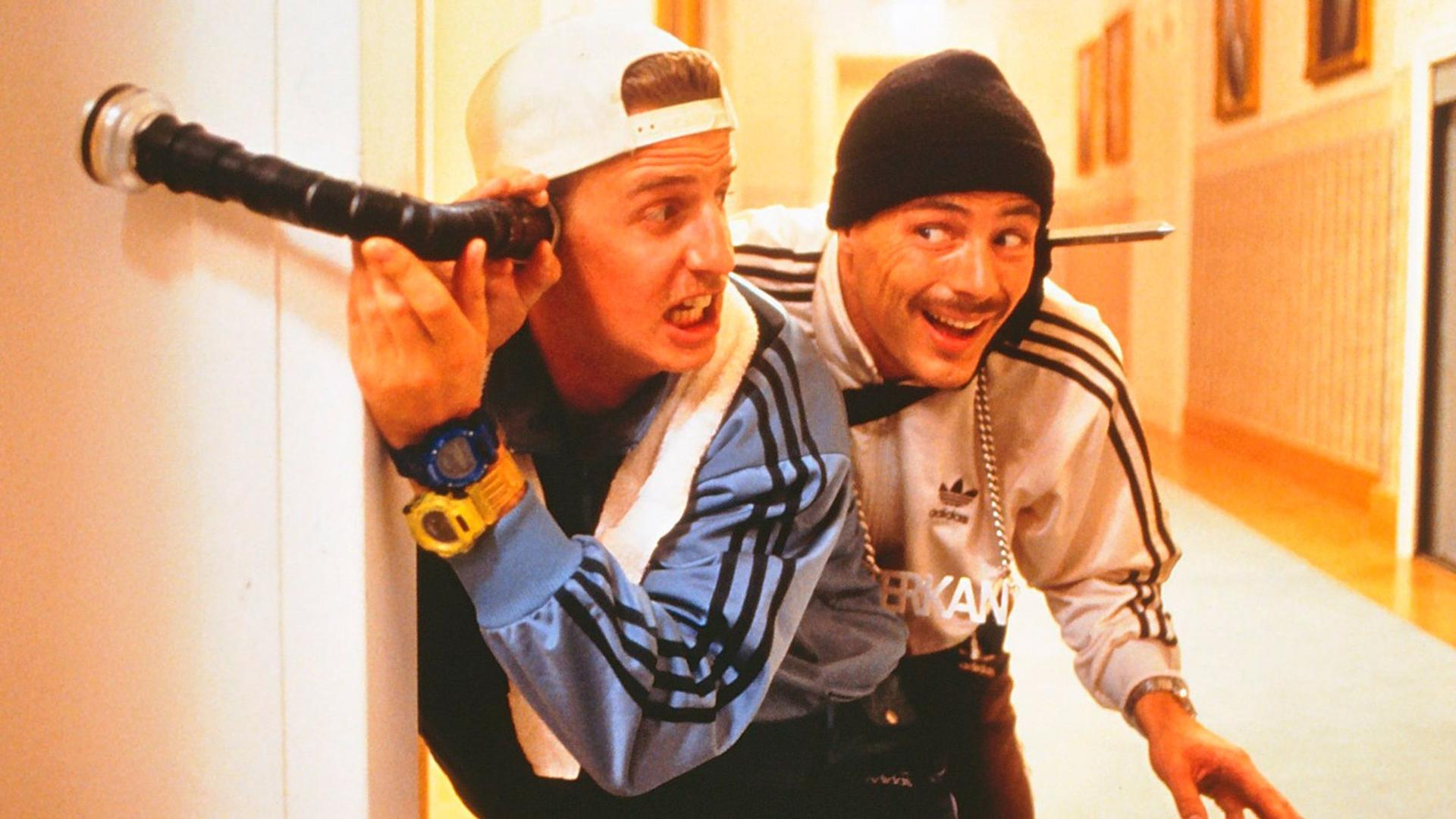 Zwei Männer in Trainingsanzügen, einer mit einem Rohr an einer Wand. Einer trägt eine Kappe und der andere eine Mütze, beide lächeln.