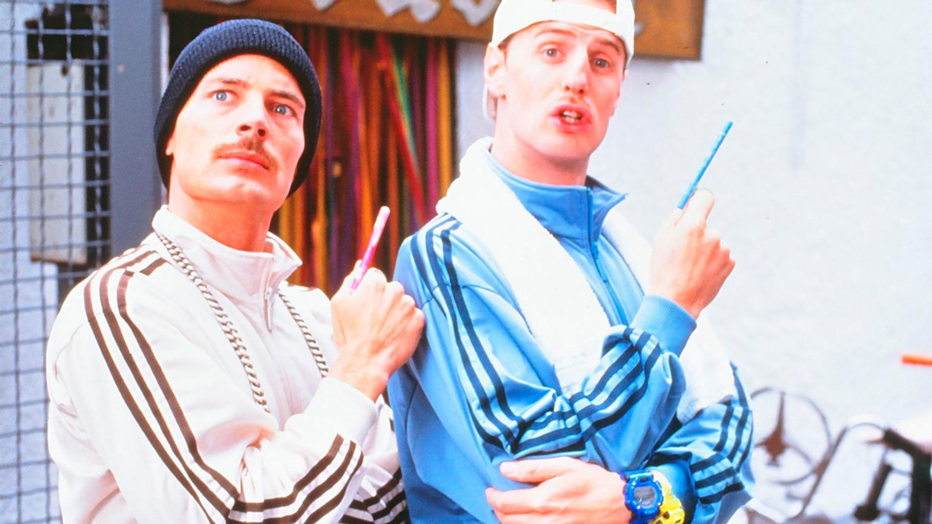 Zwei Männer in Trainingsanzügen halten Stifte. Einer trägt eine schwarze Mütze, der andere eine weiße Kappe, blauen Trainingsanzug.
