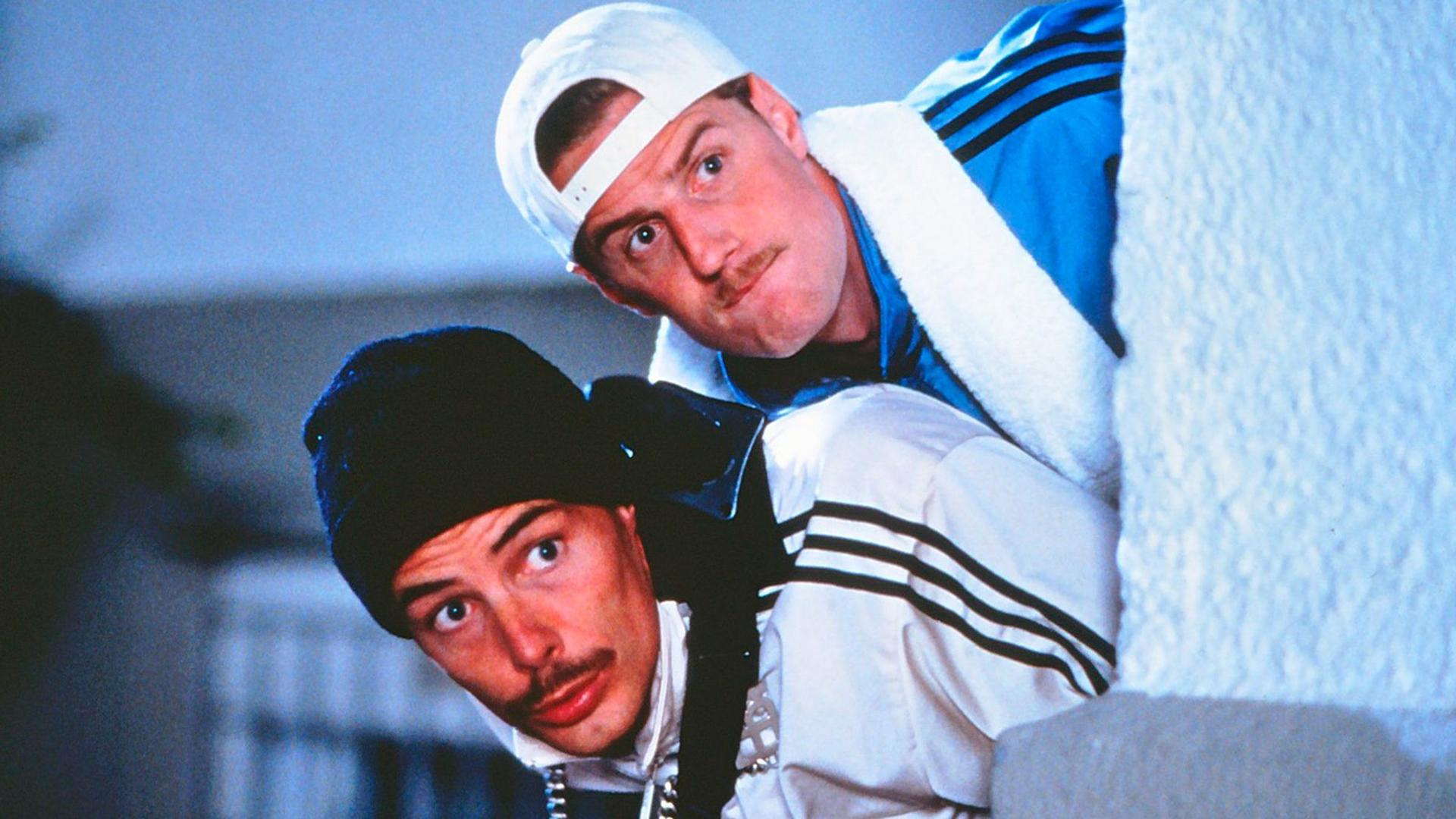 Zwei Männer, einer mit Kappe und der andere mit Mütze, spähen um eine Wand. Sie tragen Sportjacken und haben Schnurrbärte.
