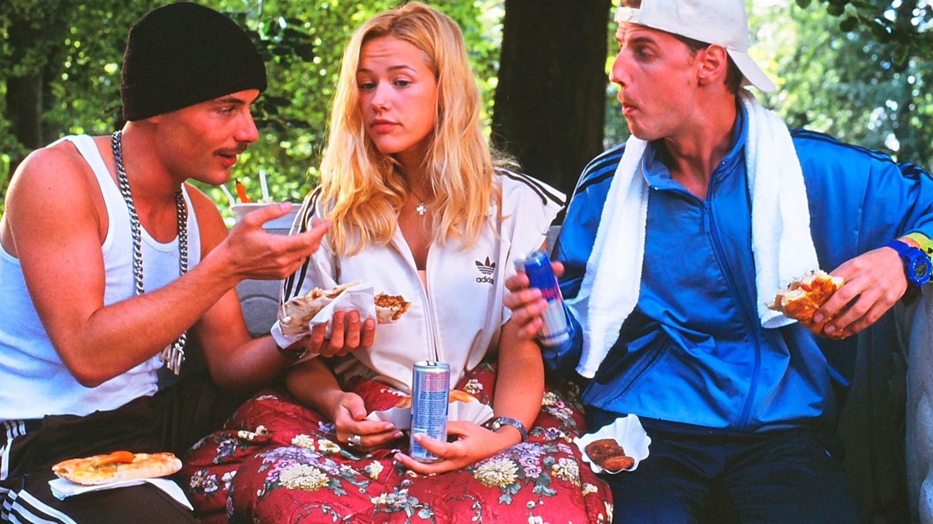 Drei Personen essen im Freien. Einer trägt eine schwarze Mütze, eine andere eine weiße Adidas-Jacke und der dritte einen blauen Trainingsanzug. Essen und Getränke sind vorhanden.