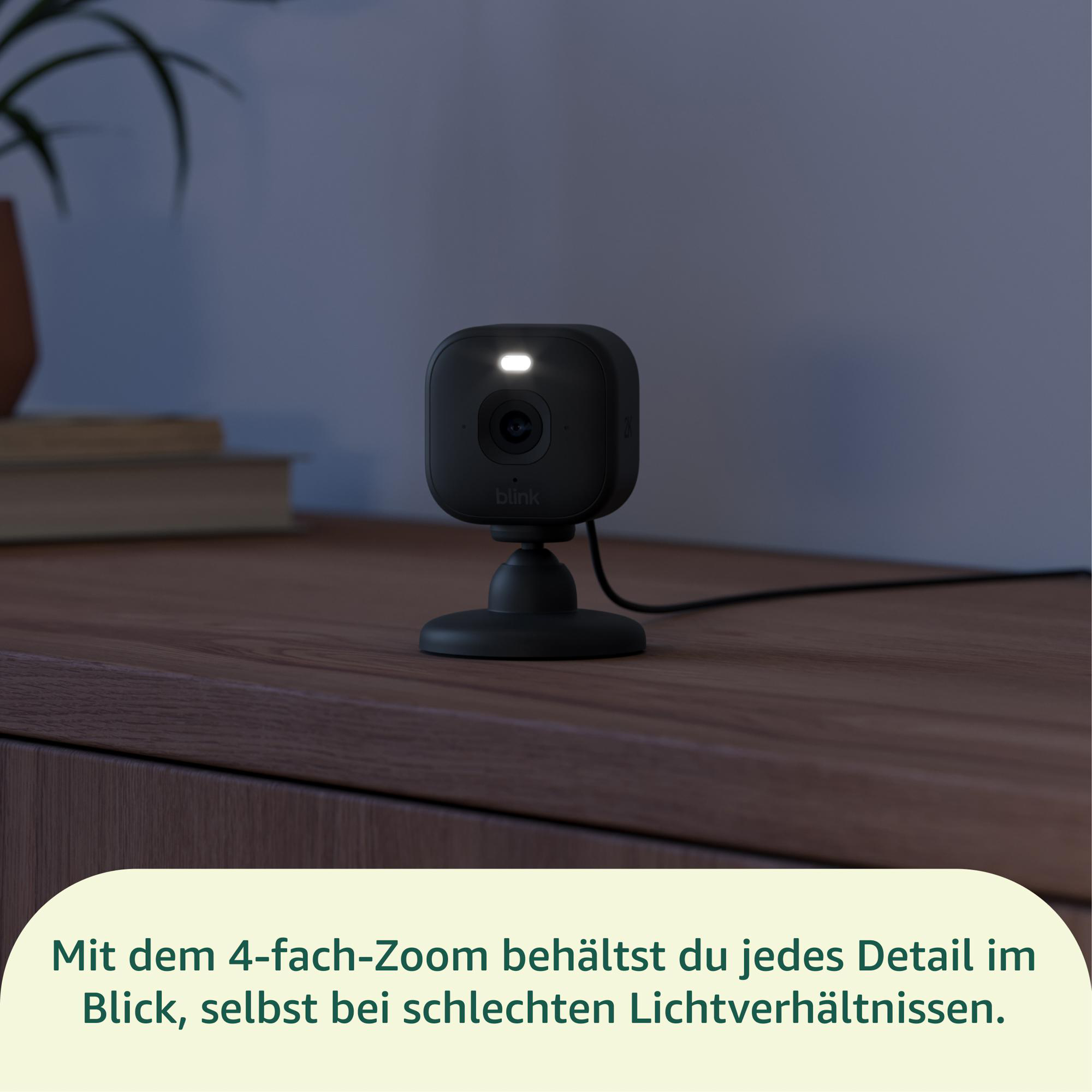Eine schwarze Blink-Sicherheitskamera mit beleuchtetem Scheinwerfer steht auf einer Holzoberfläche. Im Hintergrund ist eine Pflanze.